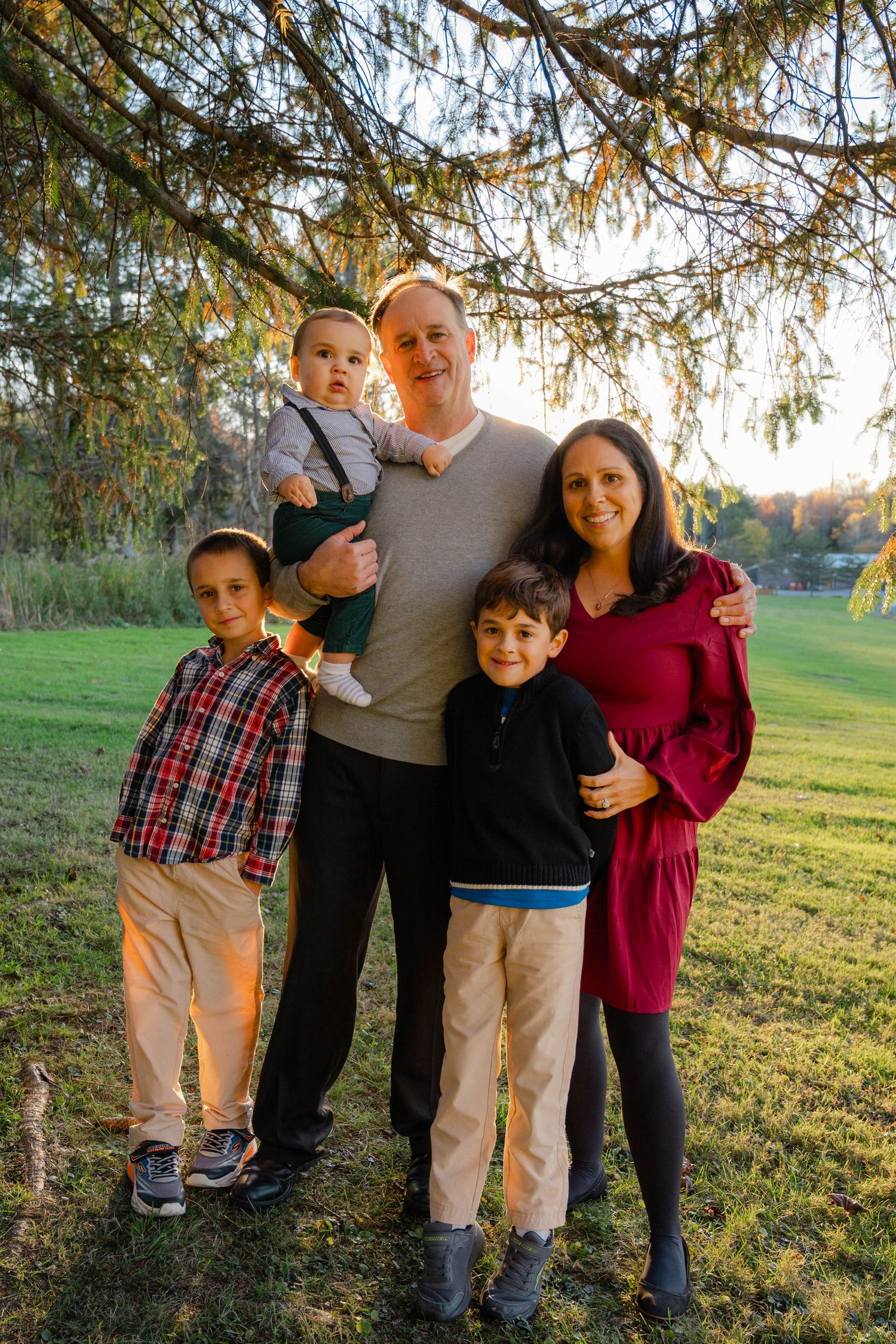 Family of five posing outdoors under pine tree in golden hour light, parents with three young boys smiling.