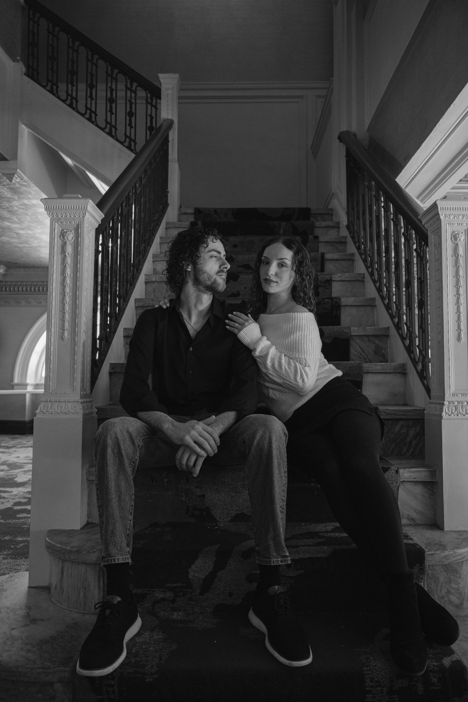 A black and white photo of a couple sitting together on grand staircase steps in an elegant home.