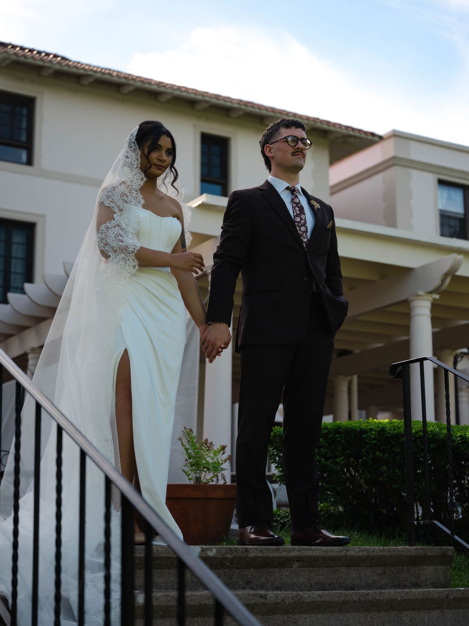 Bride and groom holding hands on steps outside wedding venue, bride wearing lace veil and slit gown.