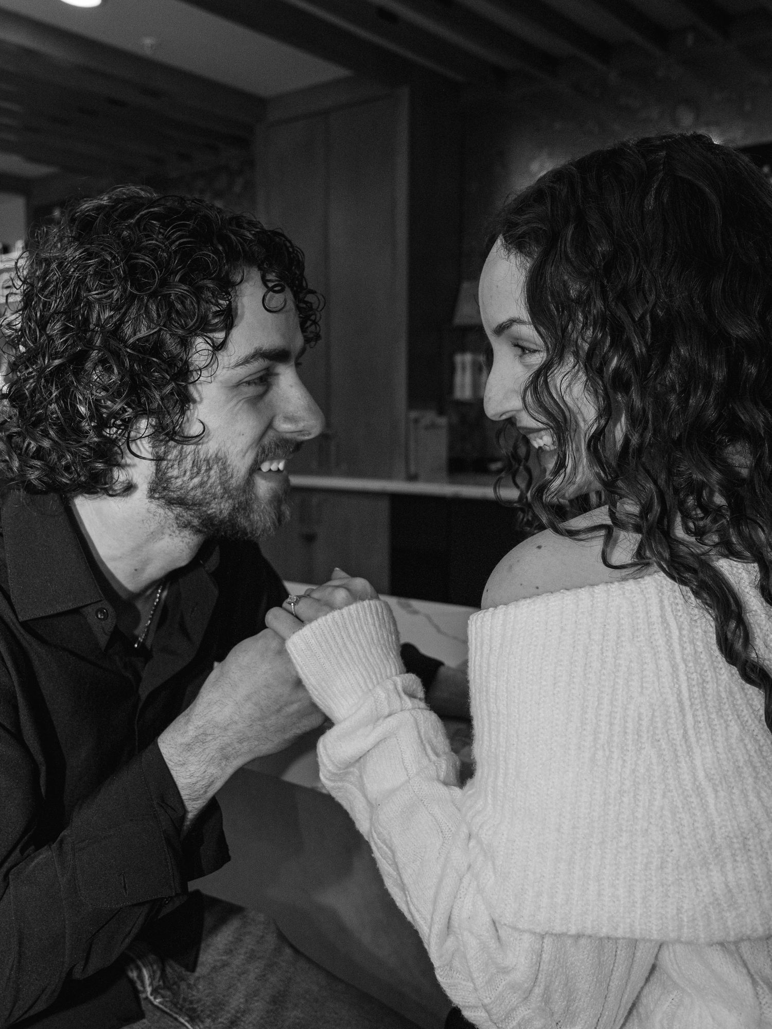 Black and white photo of a smiling couple holding hands and gazing at each other indoors.
