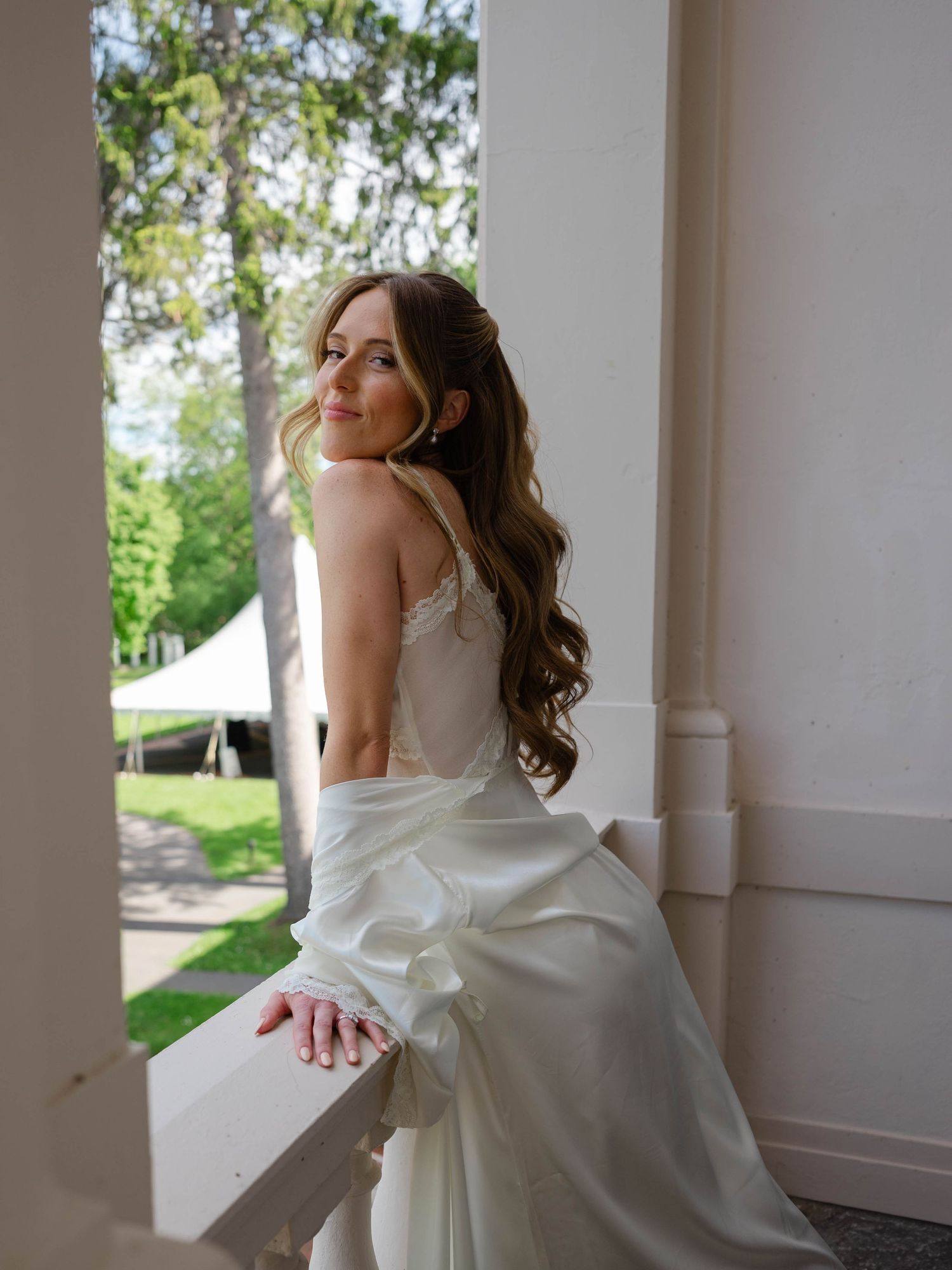 Bride in backless satin wedding gown posing on balcony with long wavy hair and delicate lace details.