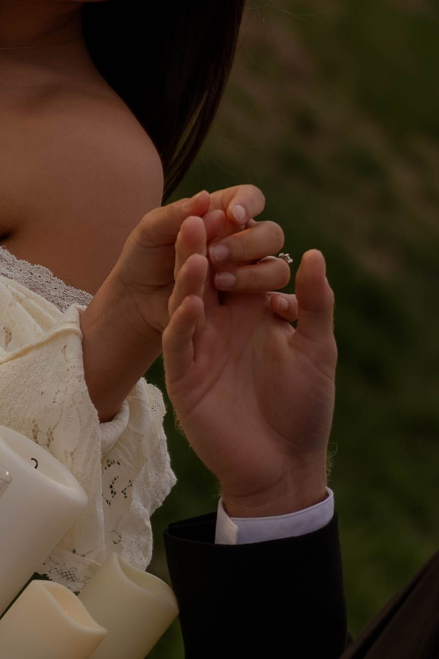 Close-up of couple holding hands with diamond engagement ring, bride in lace dress holding white bouquet.