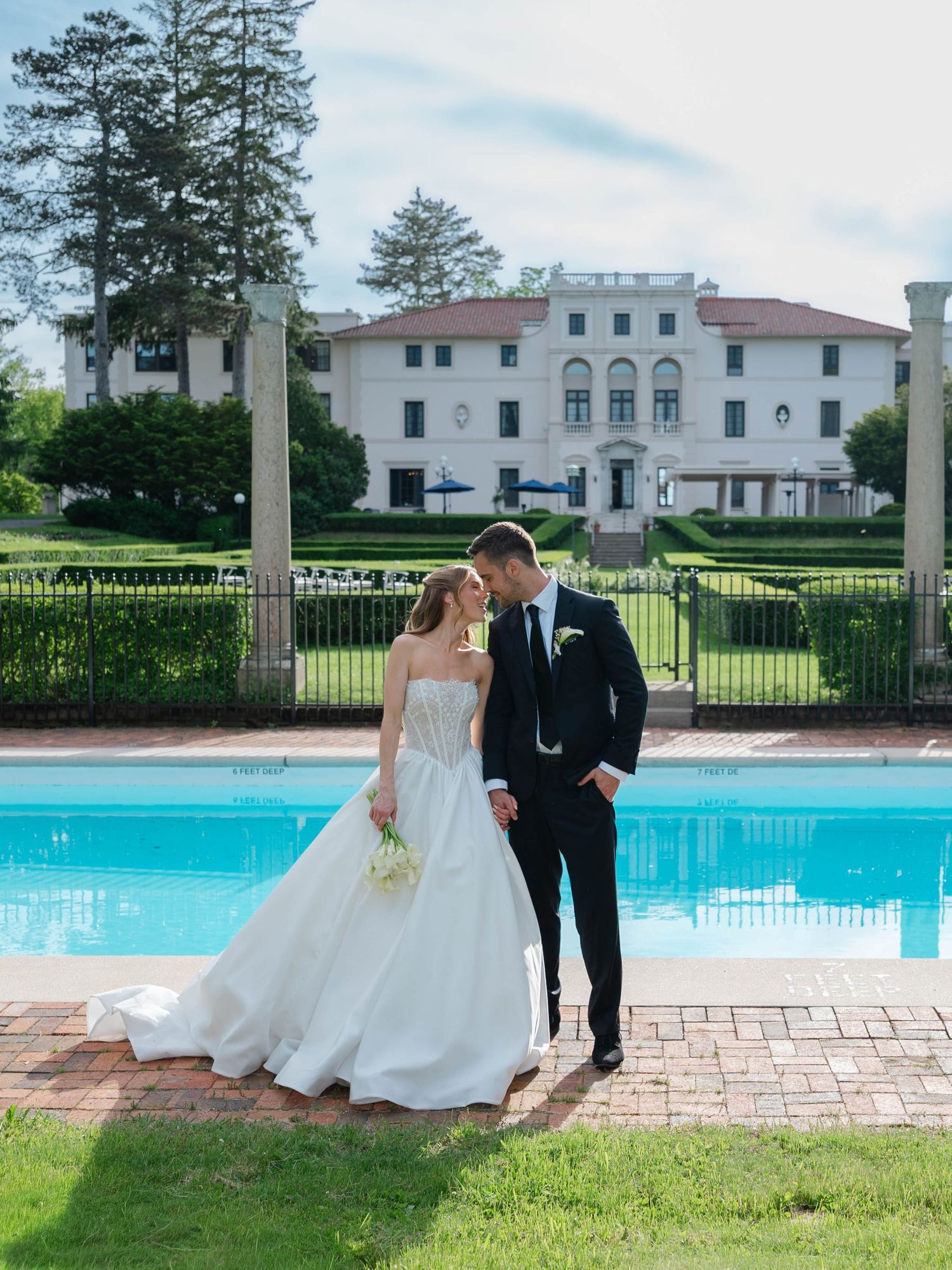 Bride and groom share tender moment by pool with grand white Italian-style mansion behind them.