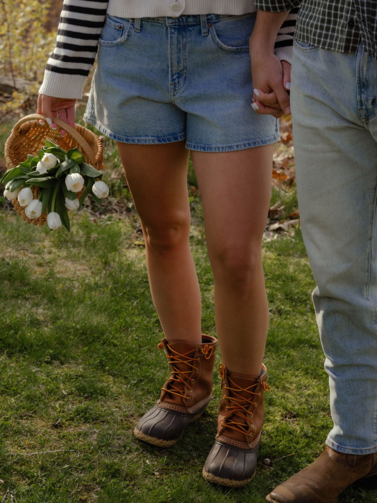 Woman in denim shorts and duck boots holding white tulips beside a man outdoors.