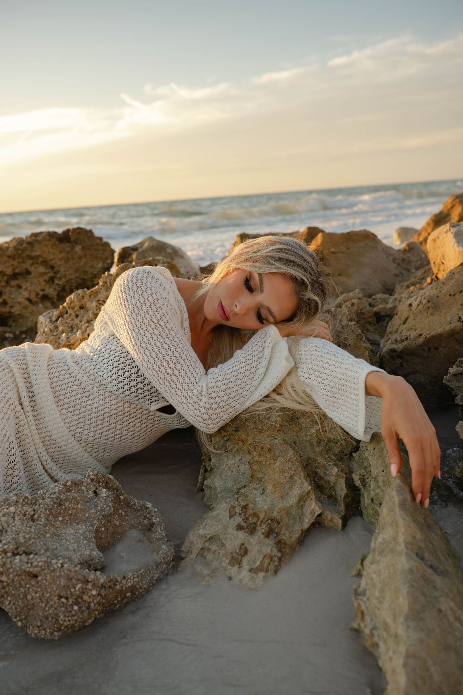 Woman in cream knit dress lying on sandy beach rocks during golden hour.