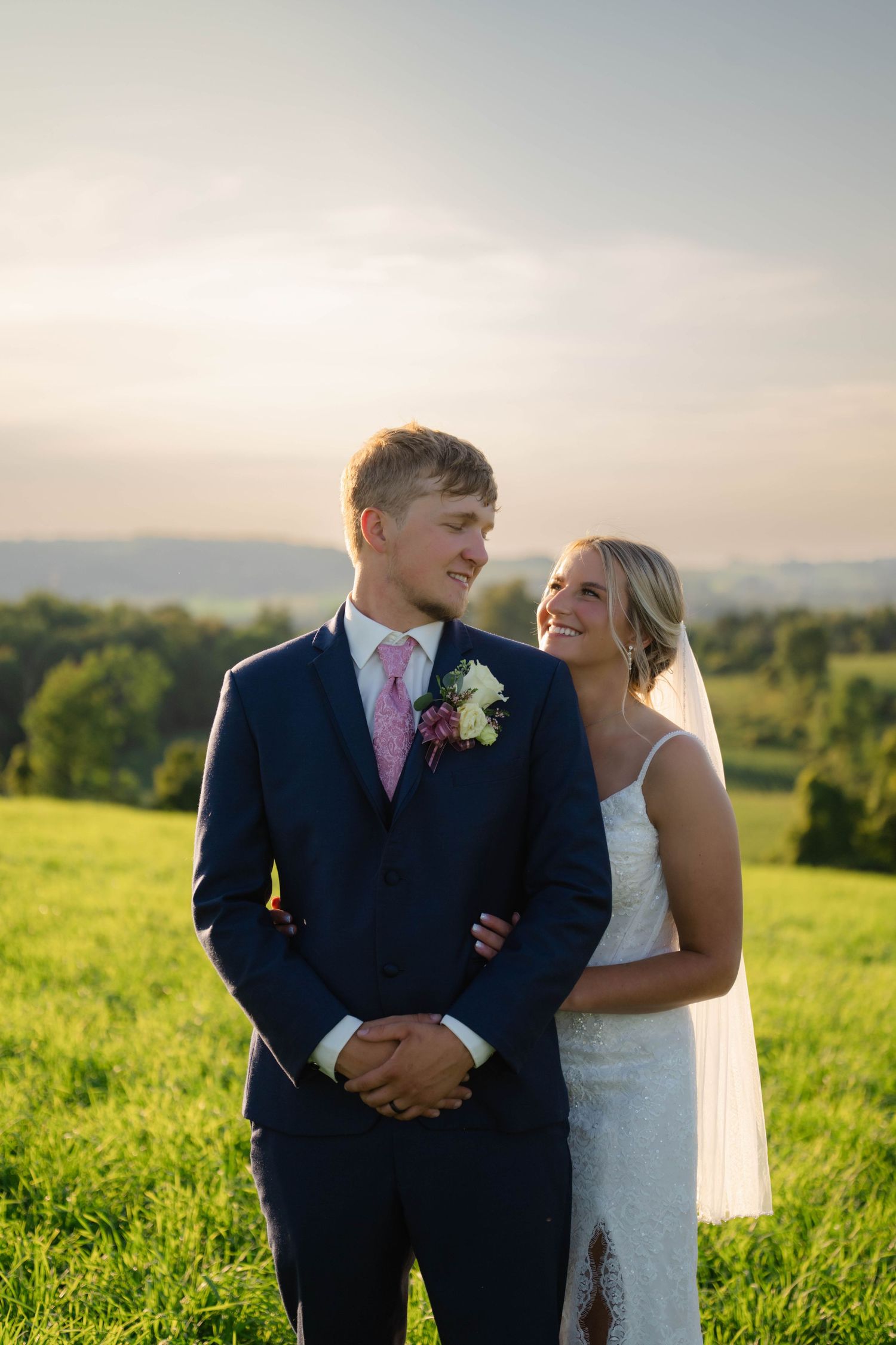A smiling bride in a white gown and veil stands with a groom in a navy suit and pink tie in a sunny green field.