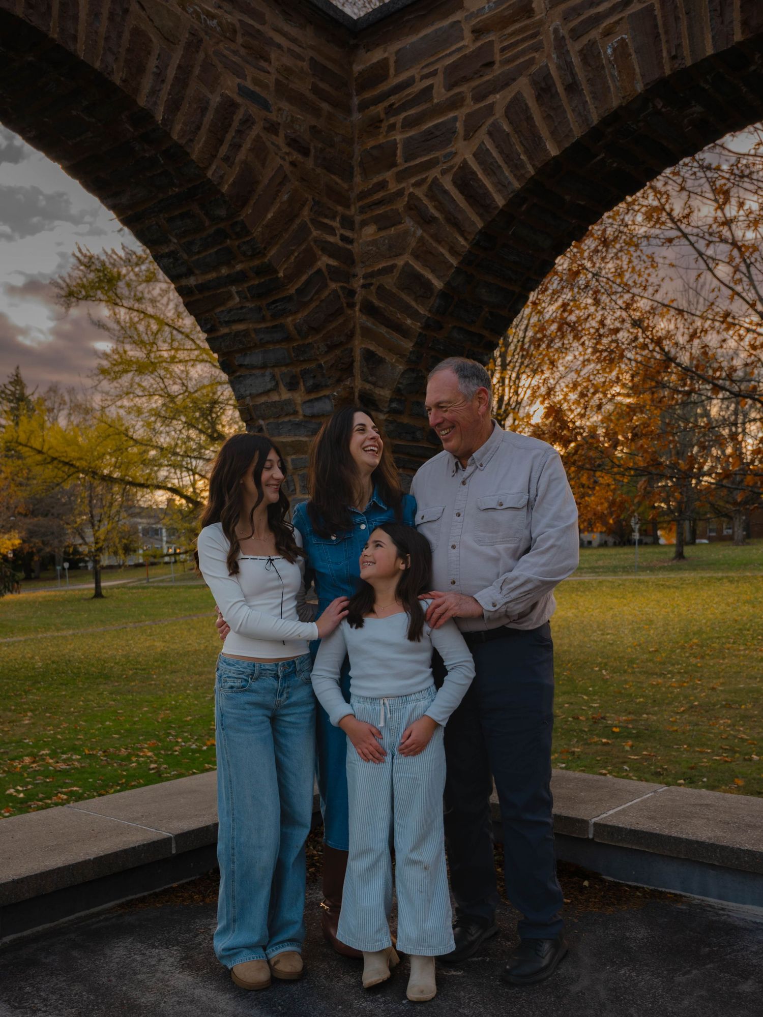Family of four smiles together under a stone arch surrounded by colorful autumn foliage at sunset.