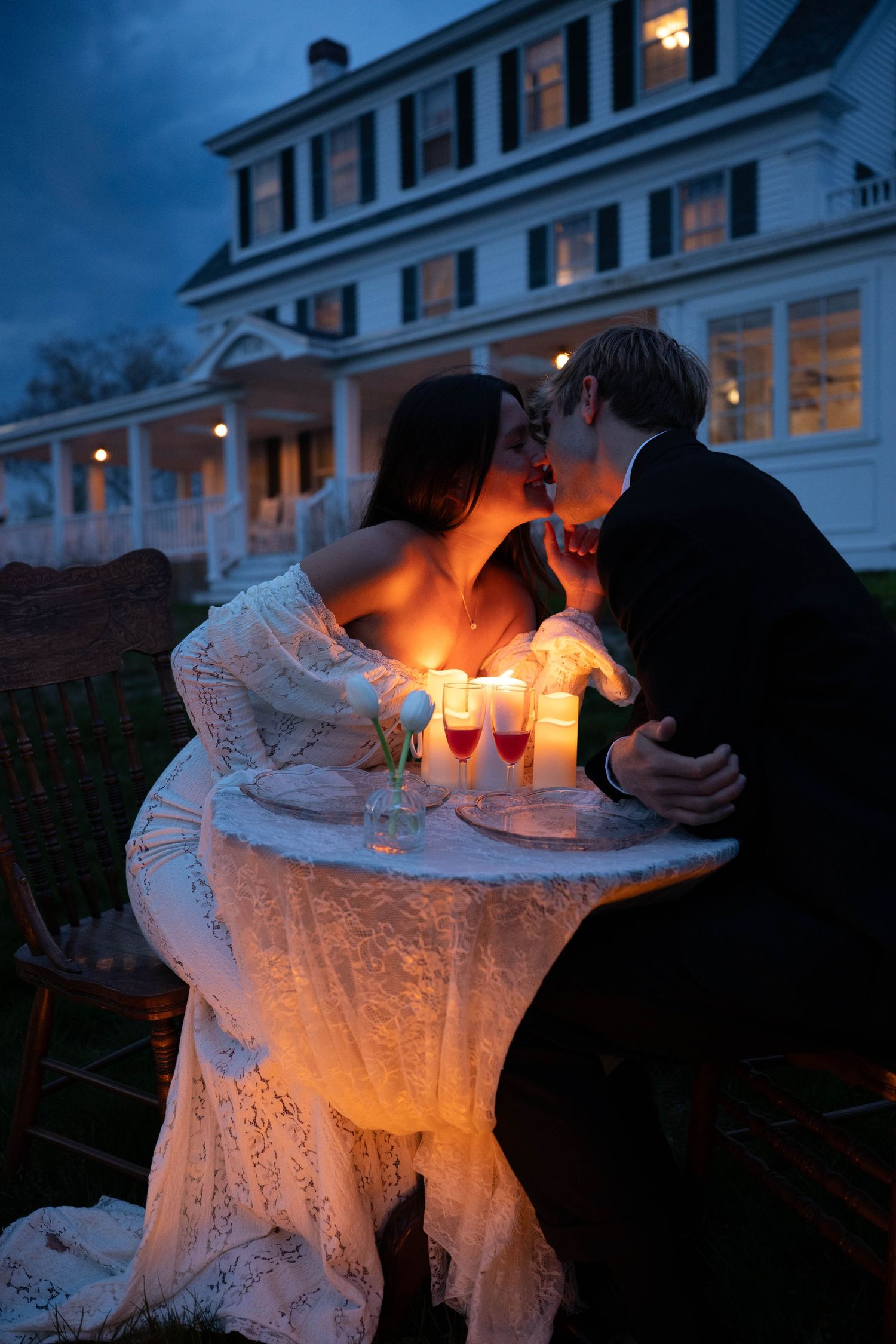 Bride and groom sharing a kiss at candlelit outdoor table at night in front of white house.