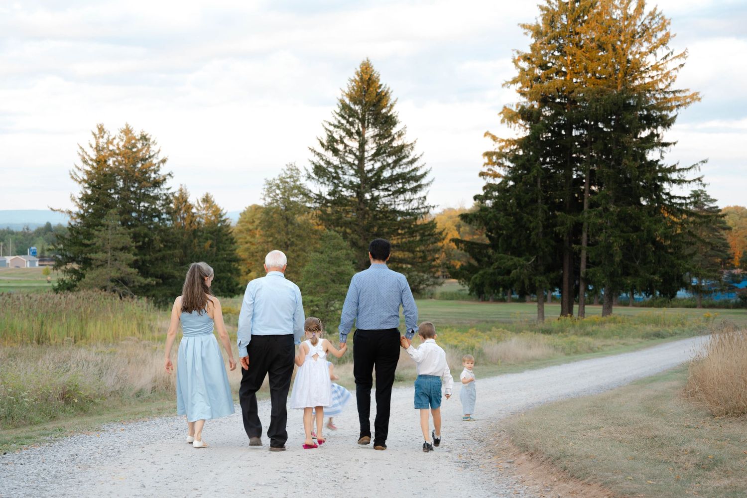 Multi-generational family walking hand-in-hand down a gravel path surrounded by tall evergreen trees in autumn.