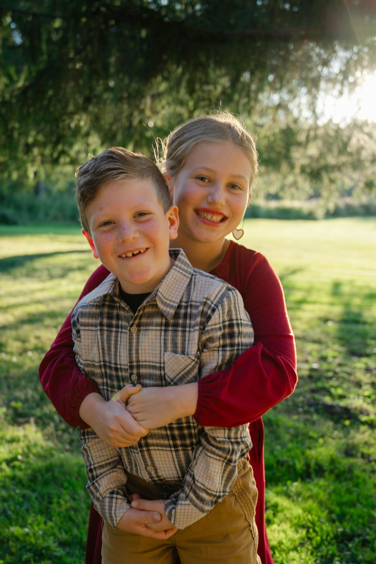 Two smiling children hugging outdoors in a sunny green park, boy in plaid shirt, girl in red top.