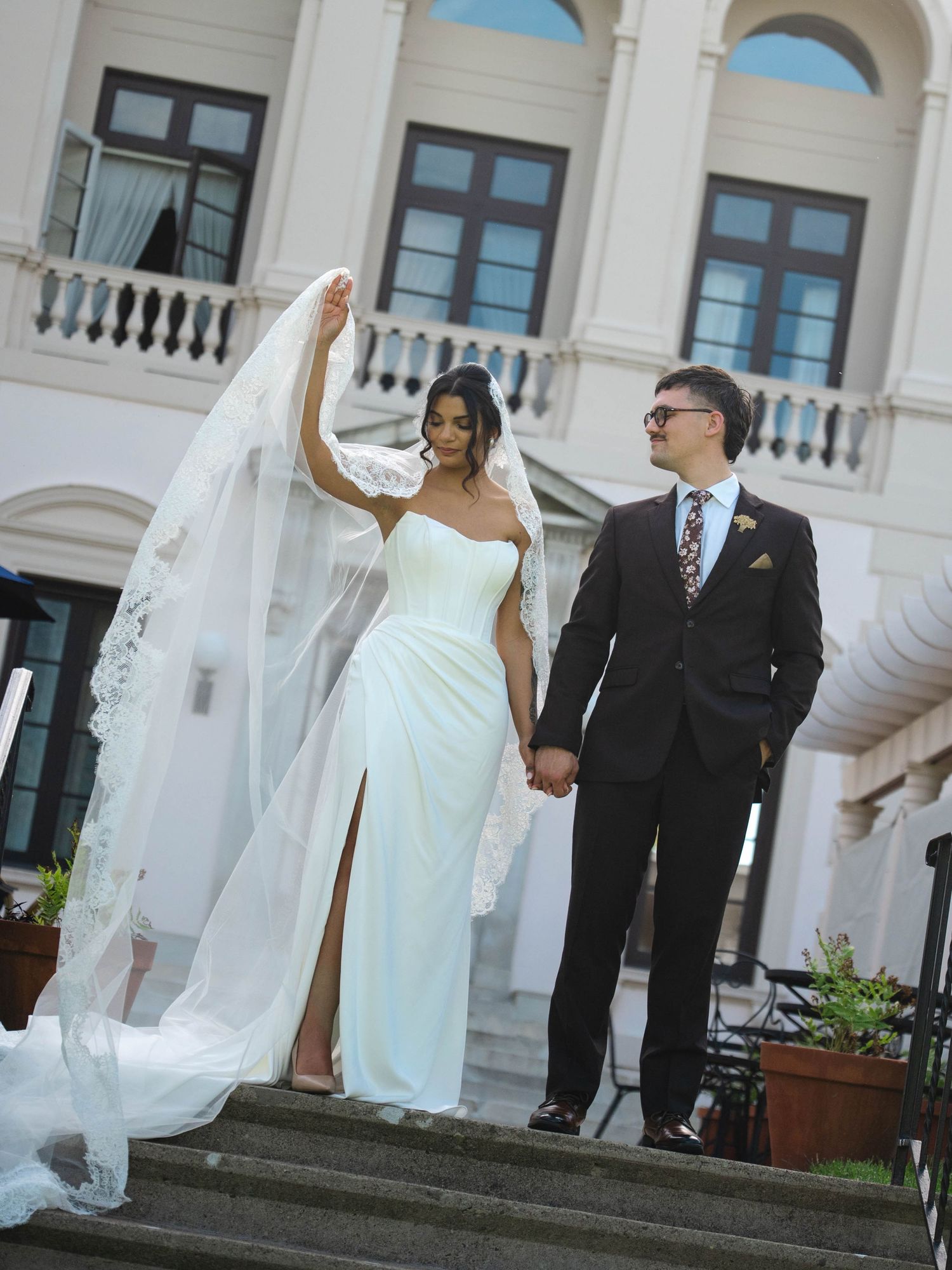Bride and groom hold hands on steps, her lace veil flowing in the wind before a classic venue.
