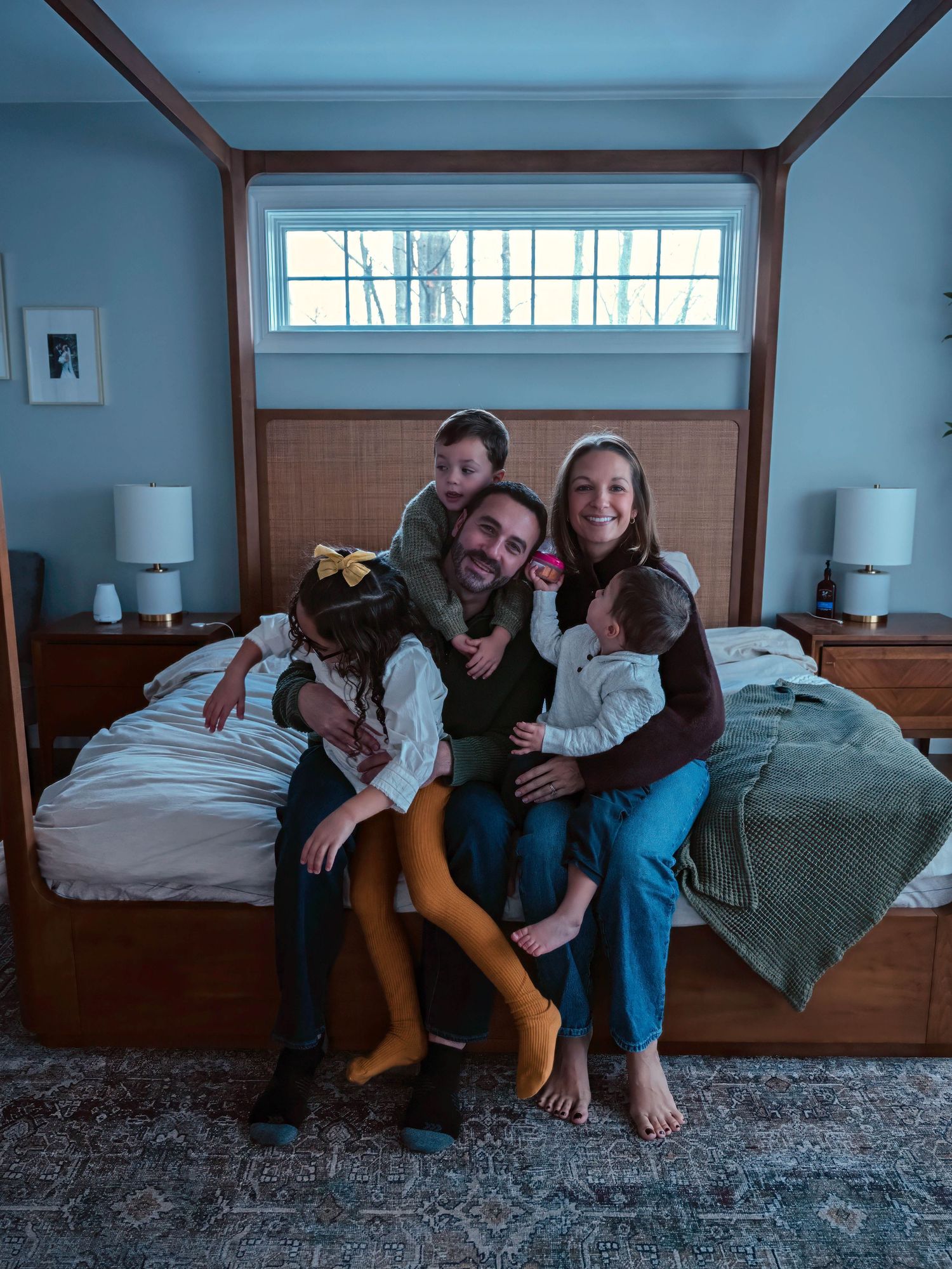 A playful family of five cuddles and laughs together on a bed in a blue bedroom.