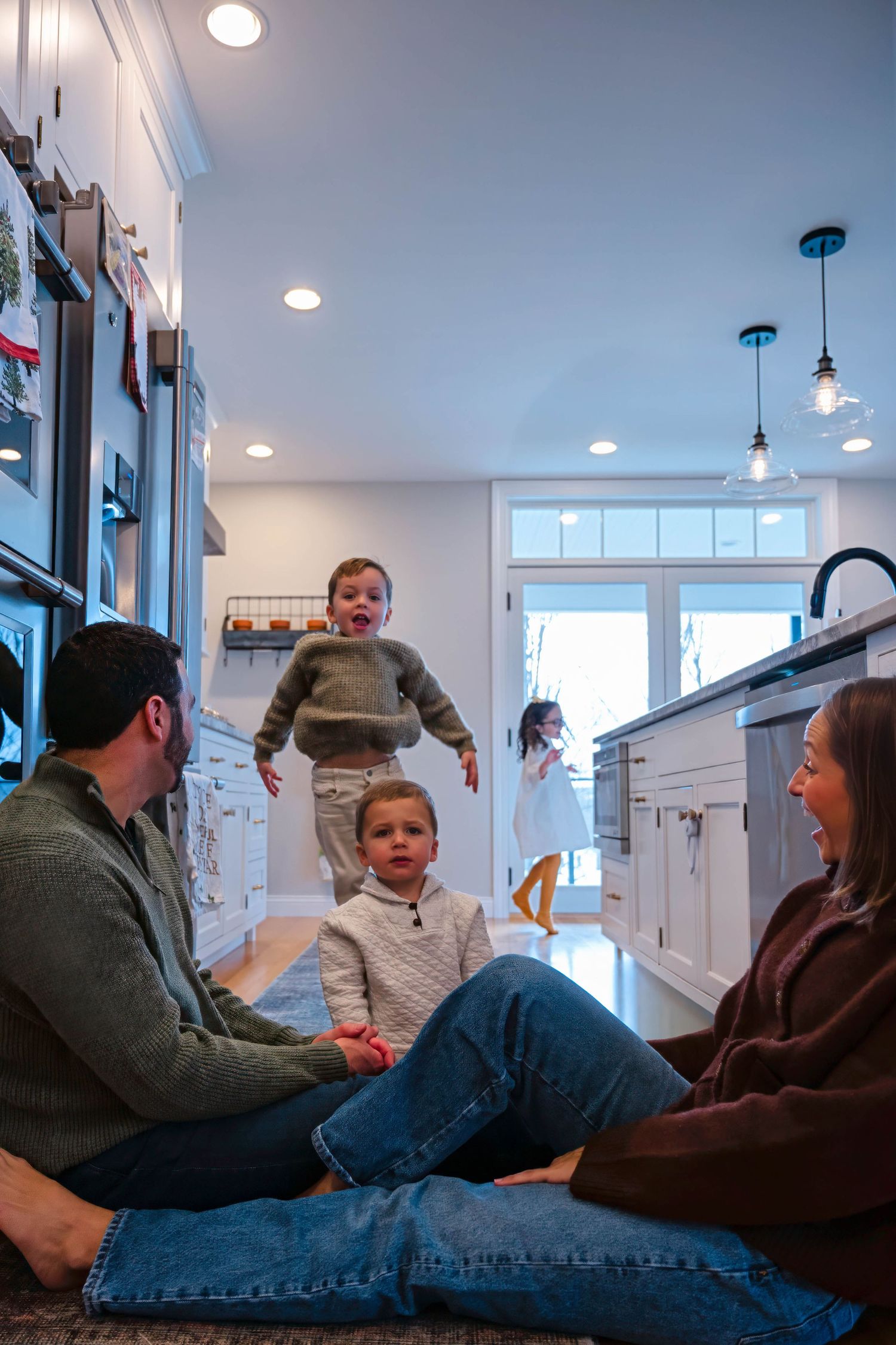 Candid family moment in modern white kitchen with toddler standing and parents sitting on floor.