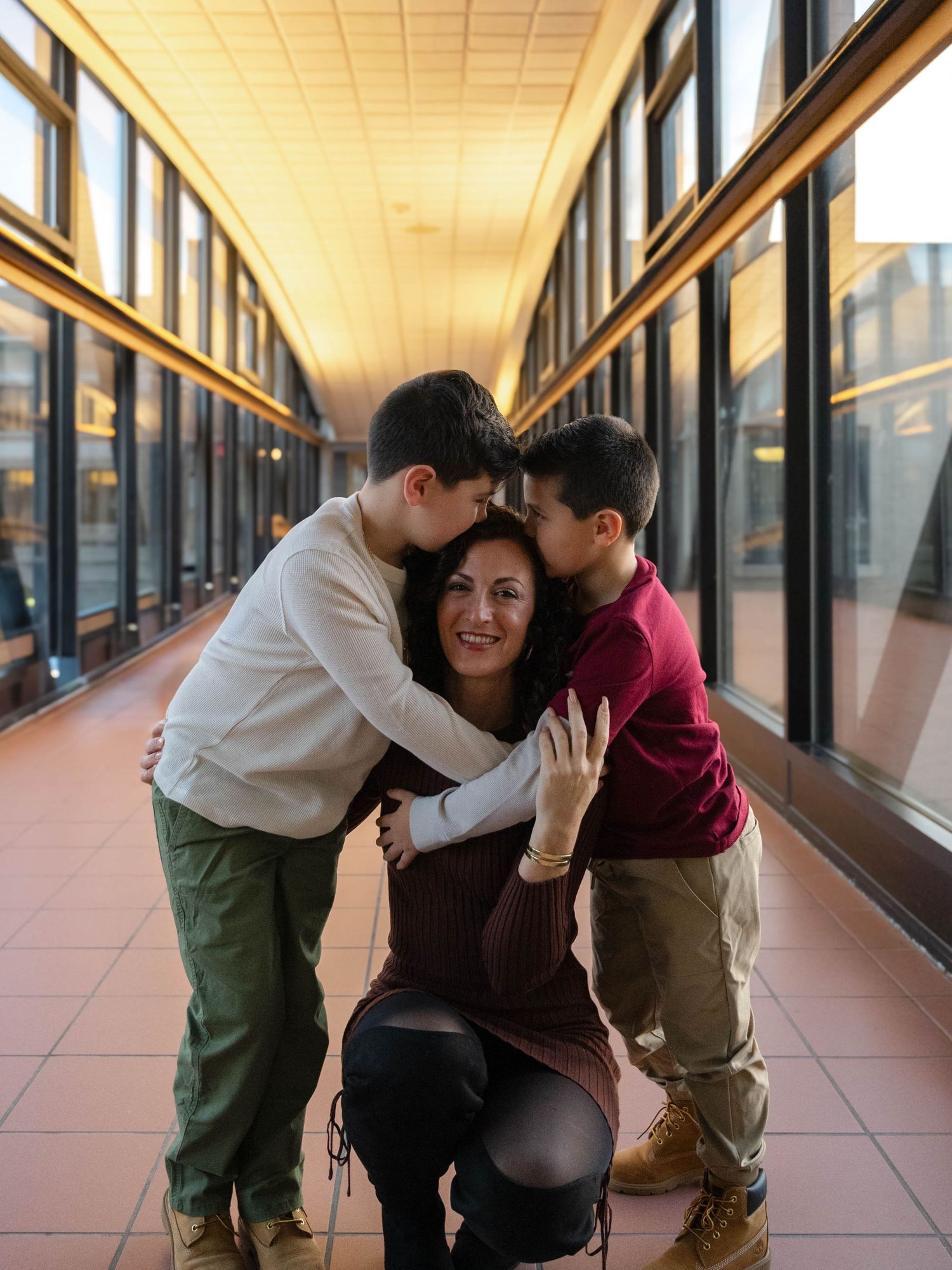 Two boys kiss their smiling pregnant mother on the cheeks in a sunlit glass corridor.