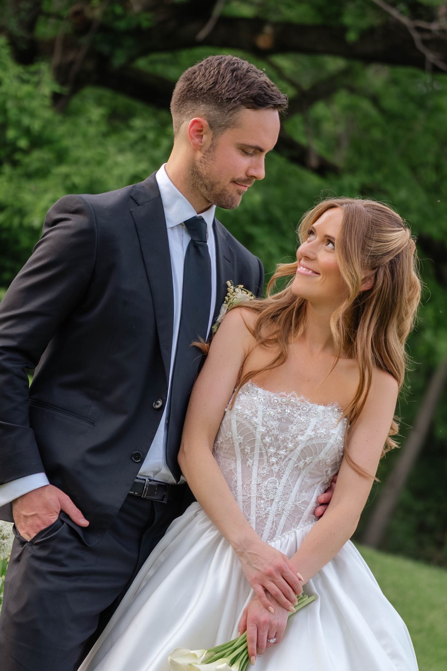 Groom in dark suit gazes down at smiling bride in strapless beaded gown holding white bouquet outdoors.