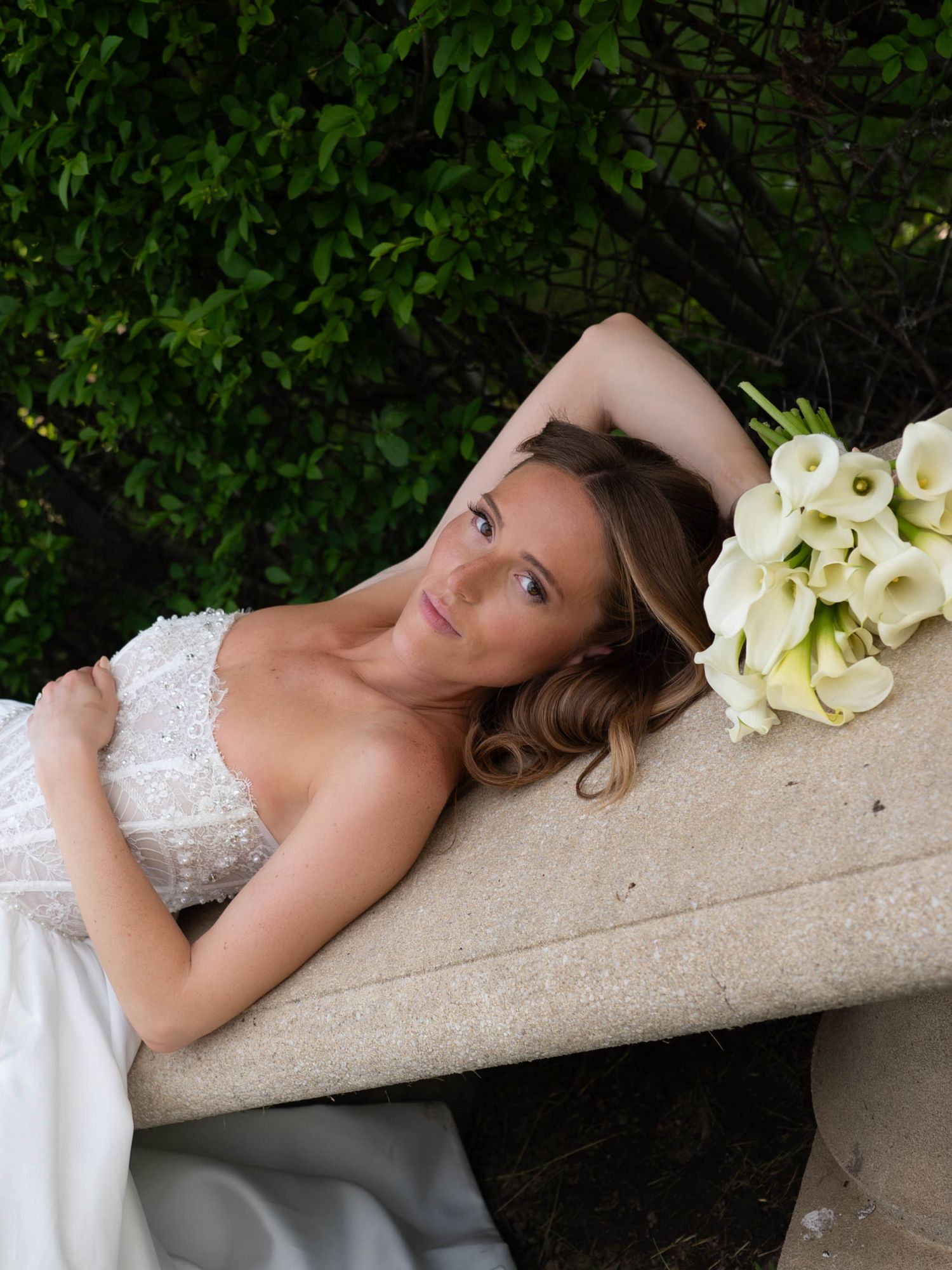 Bride in strapless lace gown reclines on a stone bench with a white calla lily bouquet beside her.