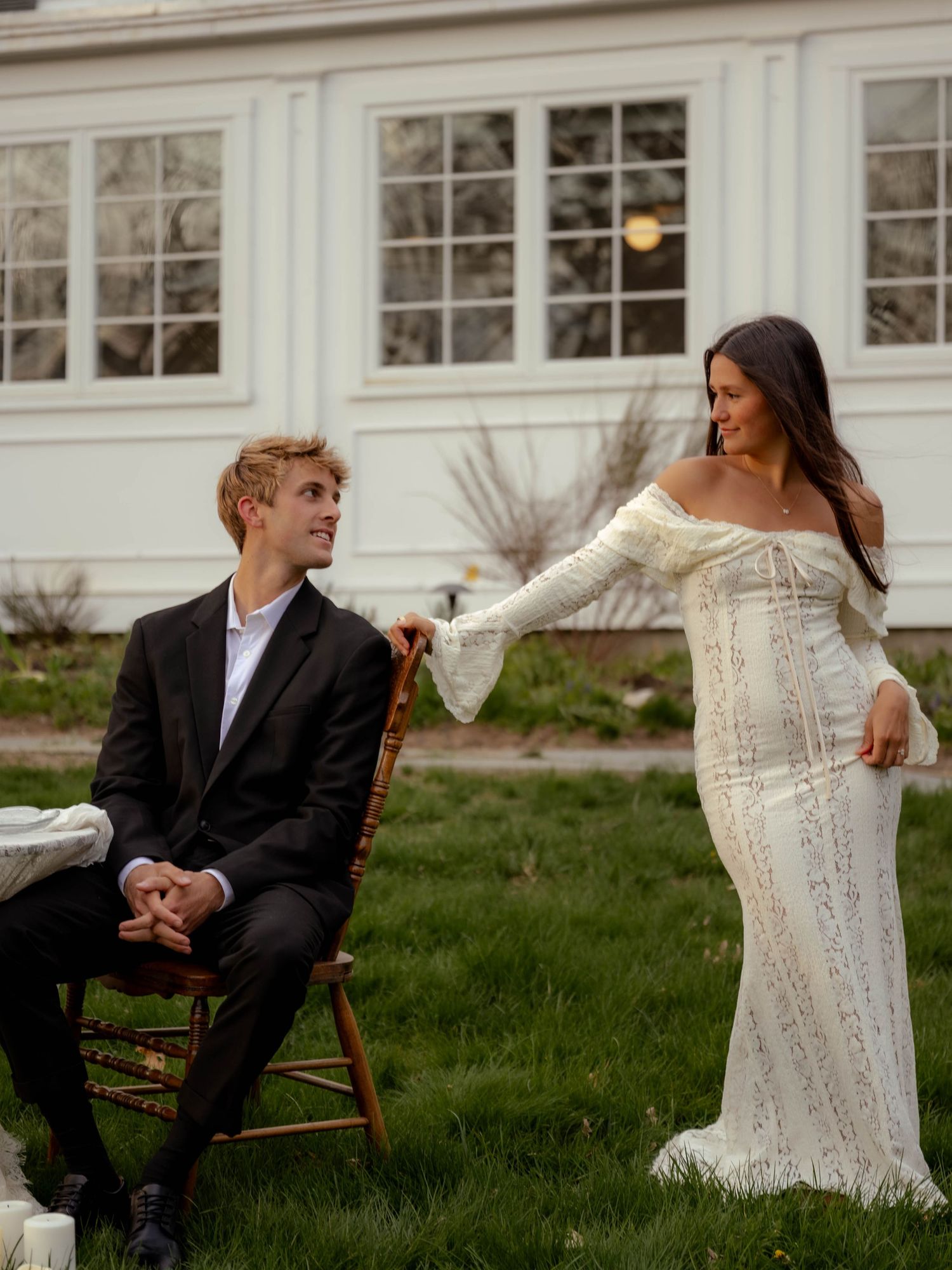 Groom seated in wooden chair smiles at bride in off-shoulder lace gown on green lawn.