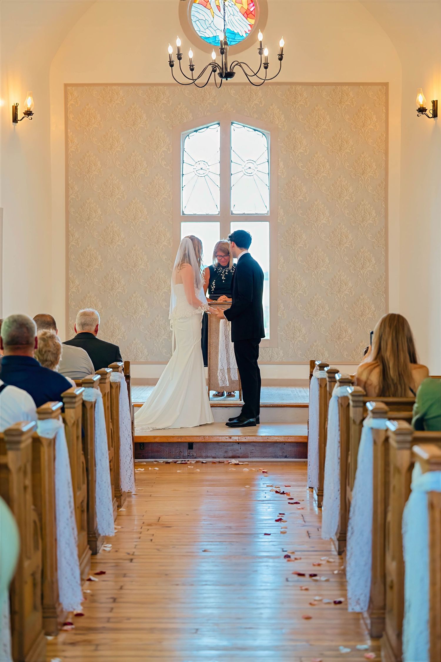 Bride and groom exchange vows at chapel altar with stained glass window, guests seated in wooden pews on hardwood floor.