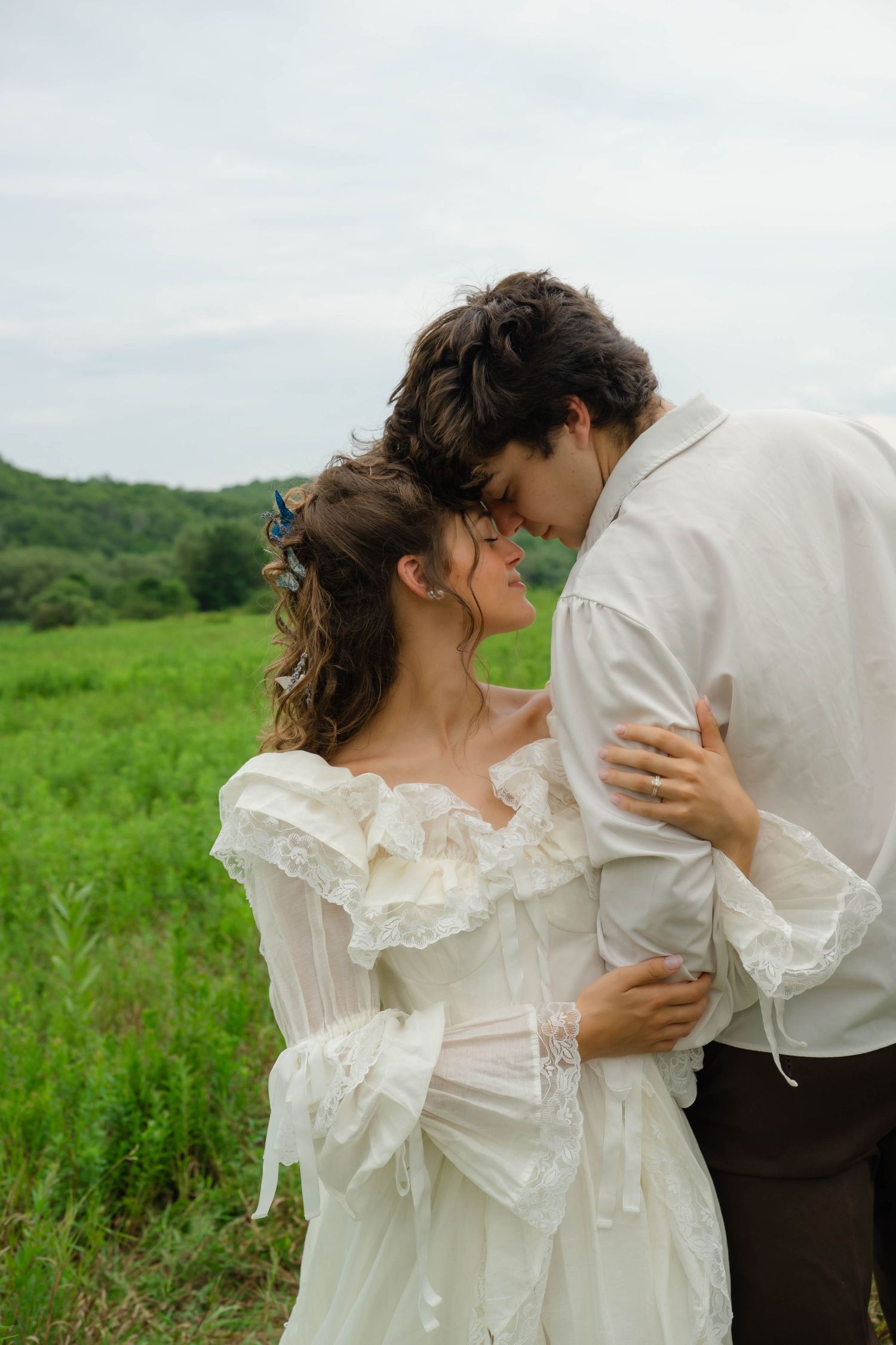 A couple in romantic attire embraces in a lush green field, the bride in a white lace dress with floral hair accessories.
