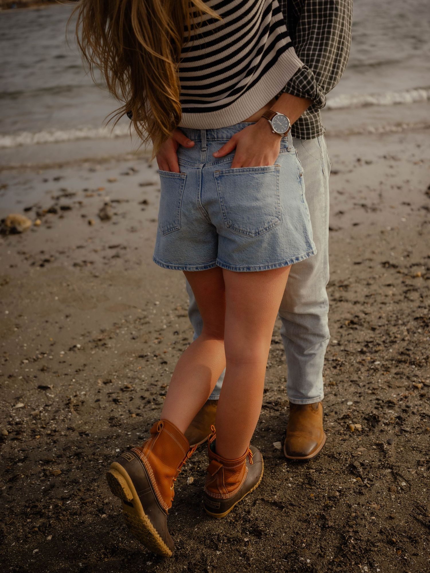 Romantic couple embracing on a sandy beach at sunset, woman wearing striped top and denim shorts.