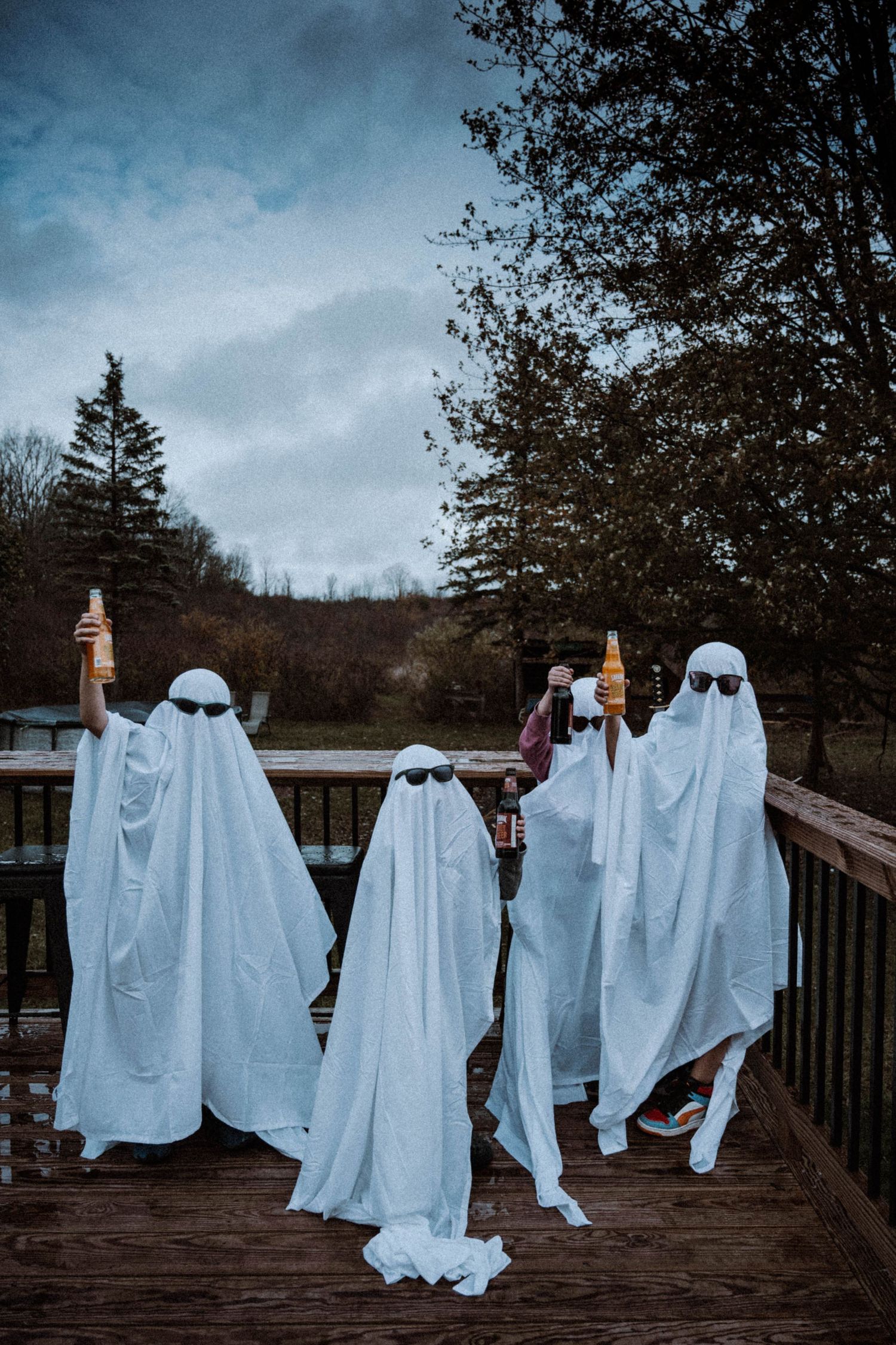 Four people in ghost costumes with sunglasses holding beer bottles on a wooden deck outdoors.