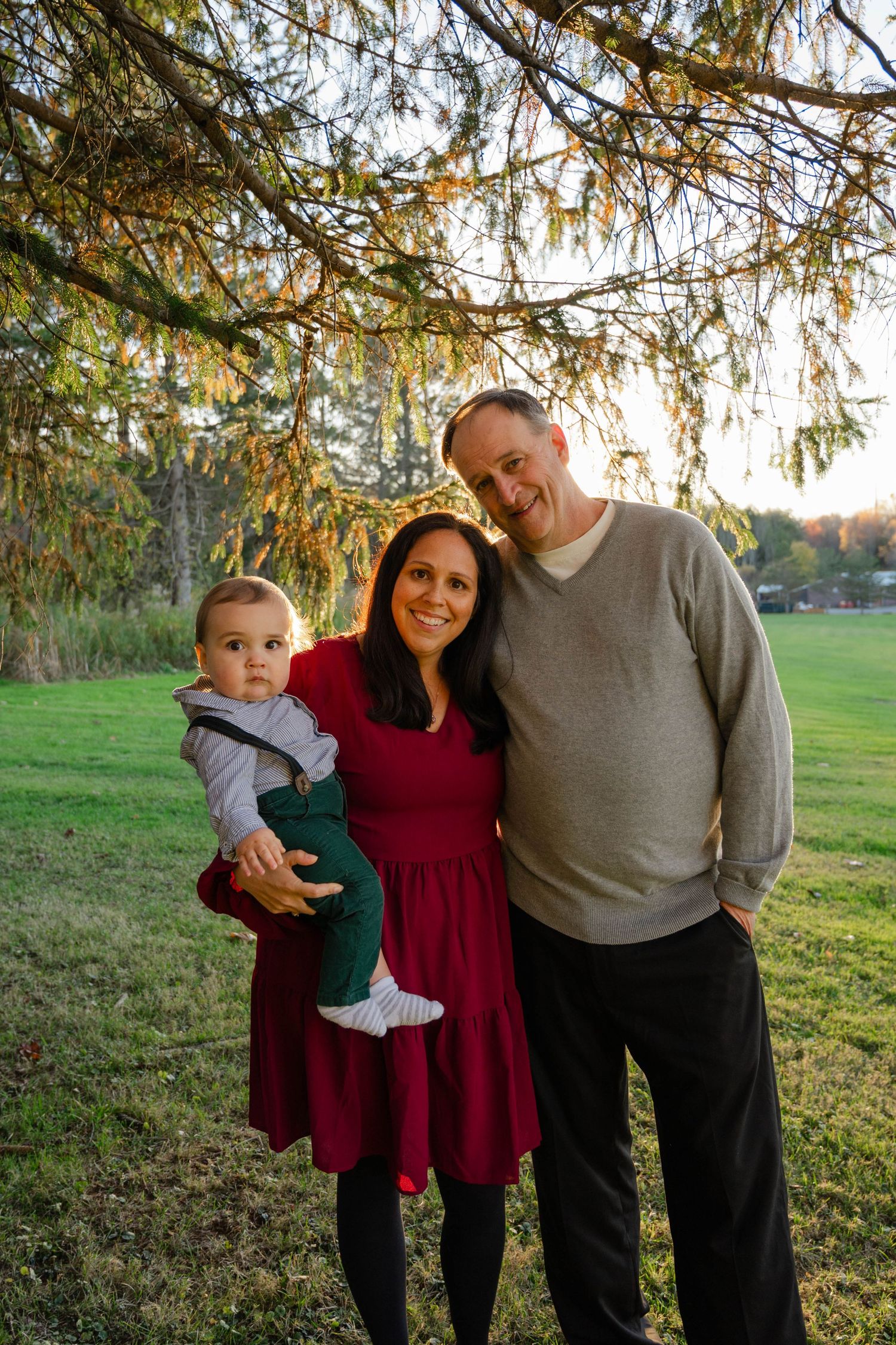 Smiling couple holding their baby boy outdoors under a pine tree in golden afternoon light.