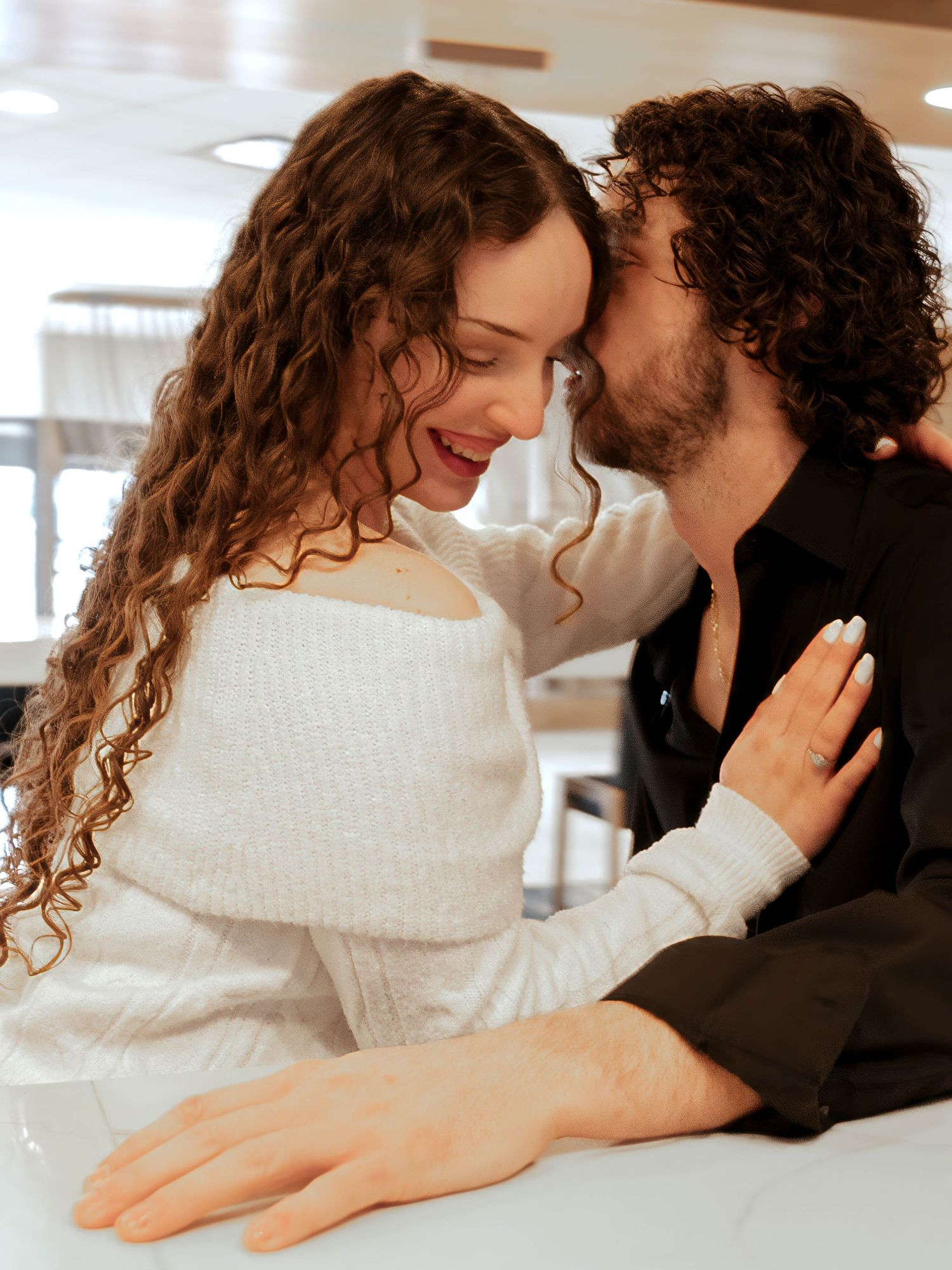 Couple sharing a tender kiss at a white counter, woman in off-shoulder sweater smiling.