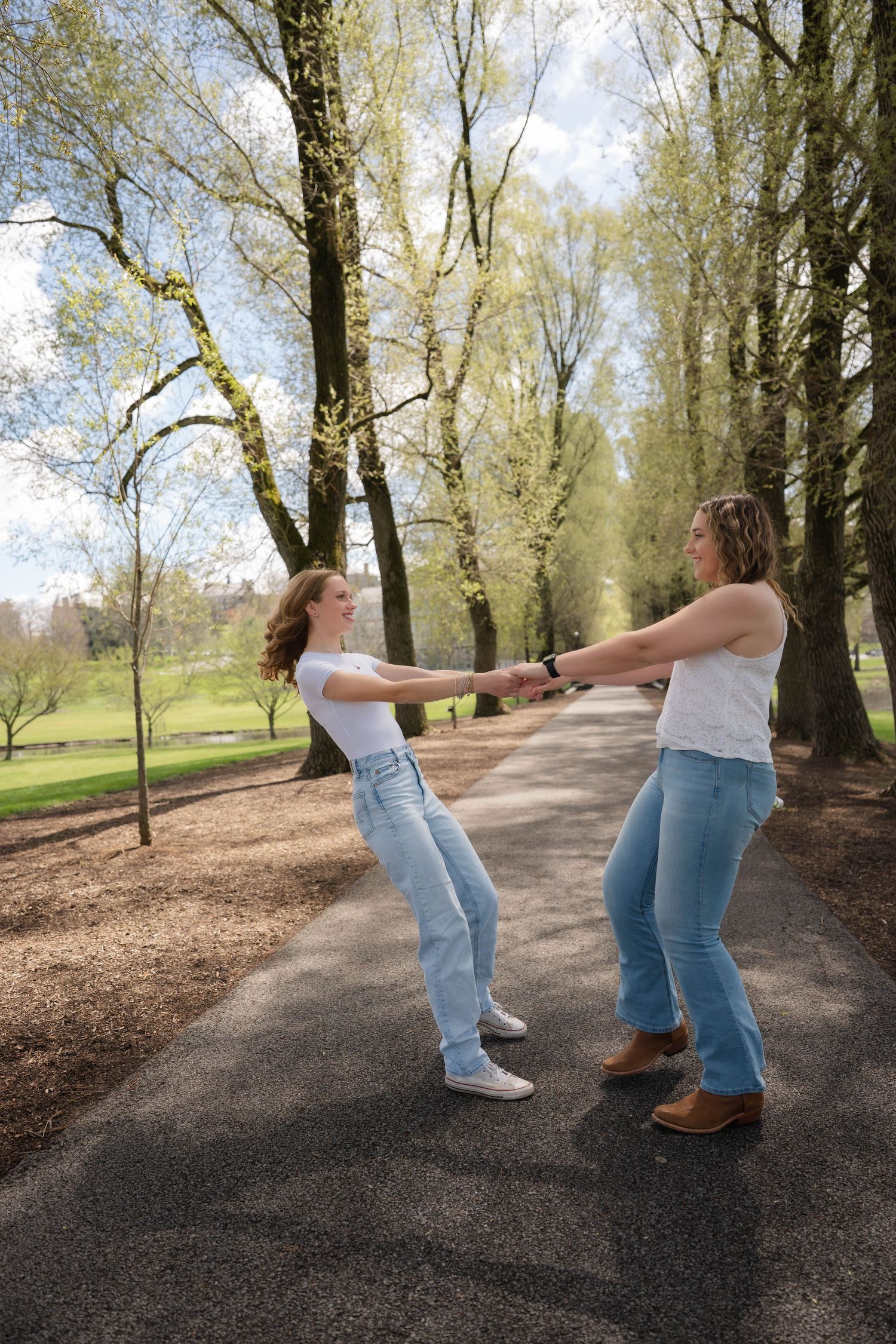 Two young women in matching white tops and jeans playfully spinning each other on a park path.