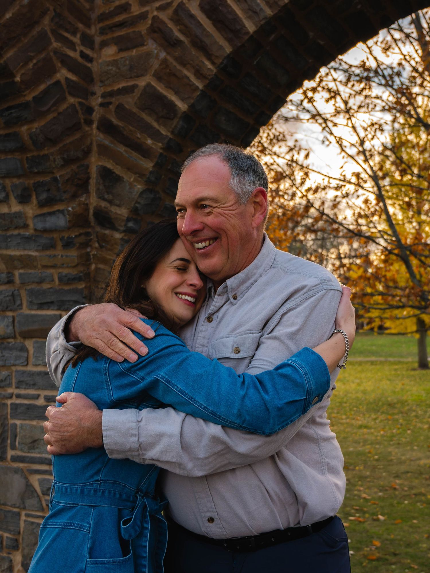 Smiling woman in blue denim dress hugs a laughing older man under a rustic stone arch outdoors.
