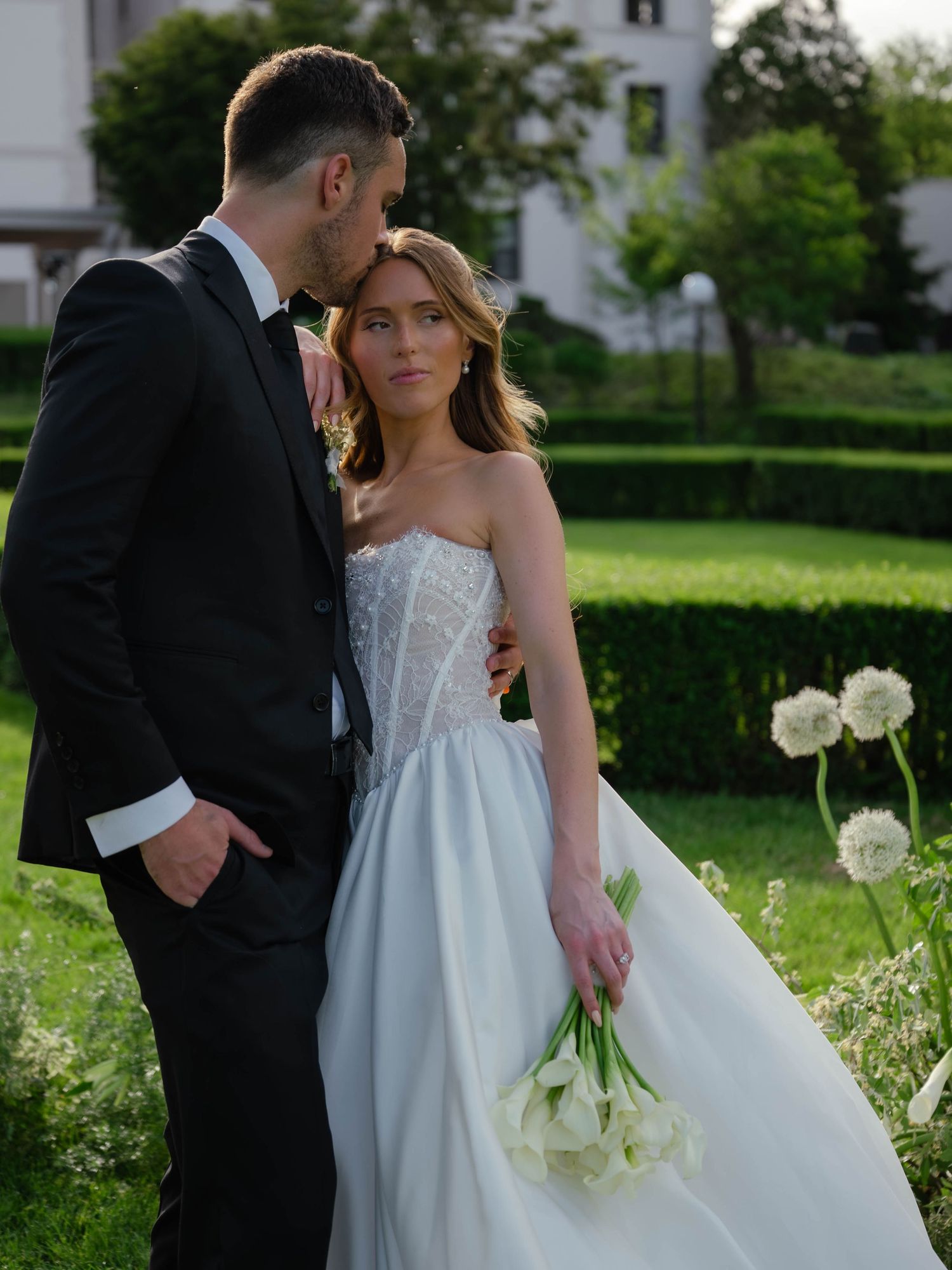 Bride in strapless corset wedding gown holding white tulips with groom in black suit in garden.