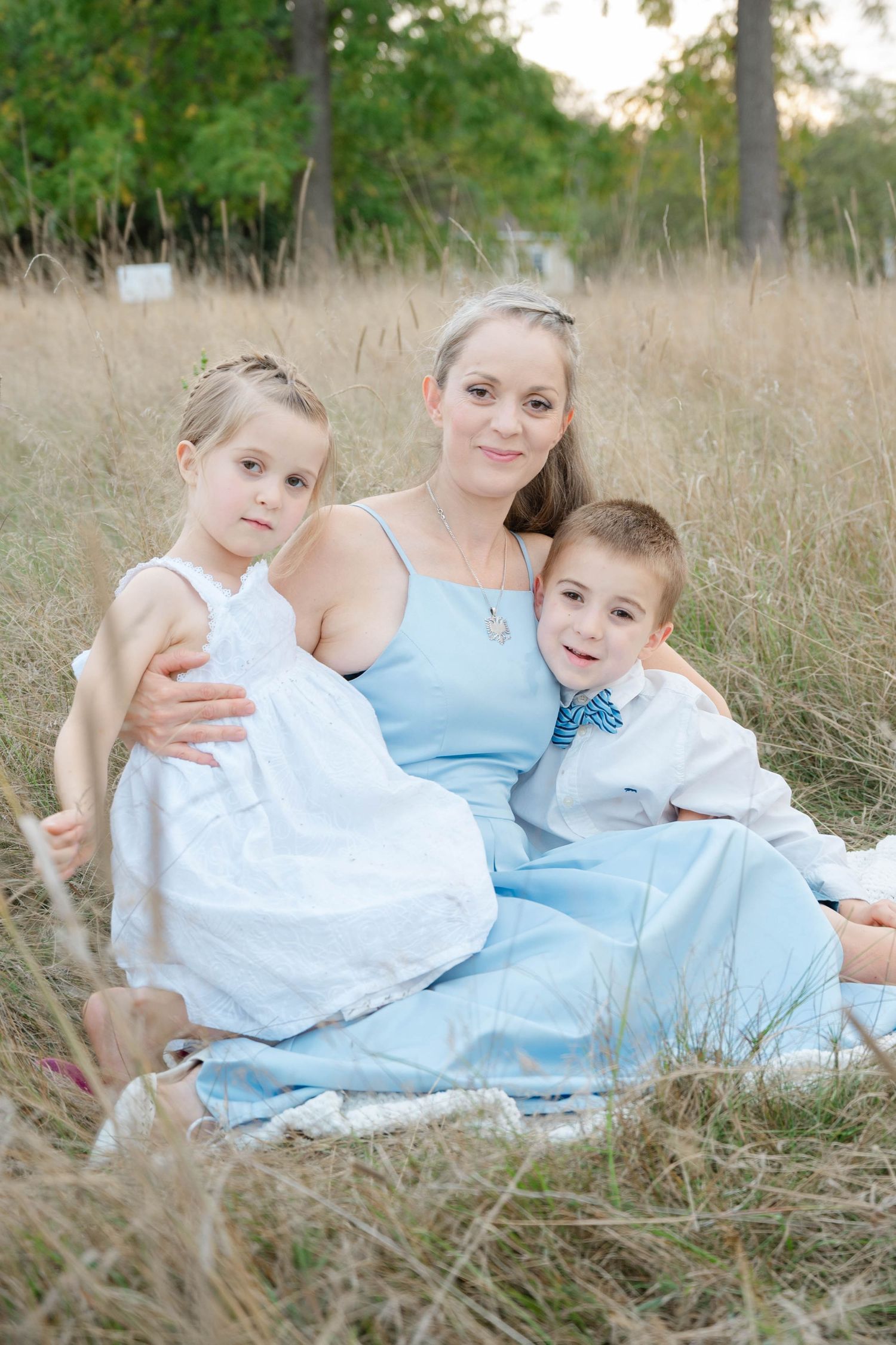 Smiling mother in blue dress sitting outdoors in a golden field with her young daughter and son on a blanket.