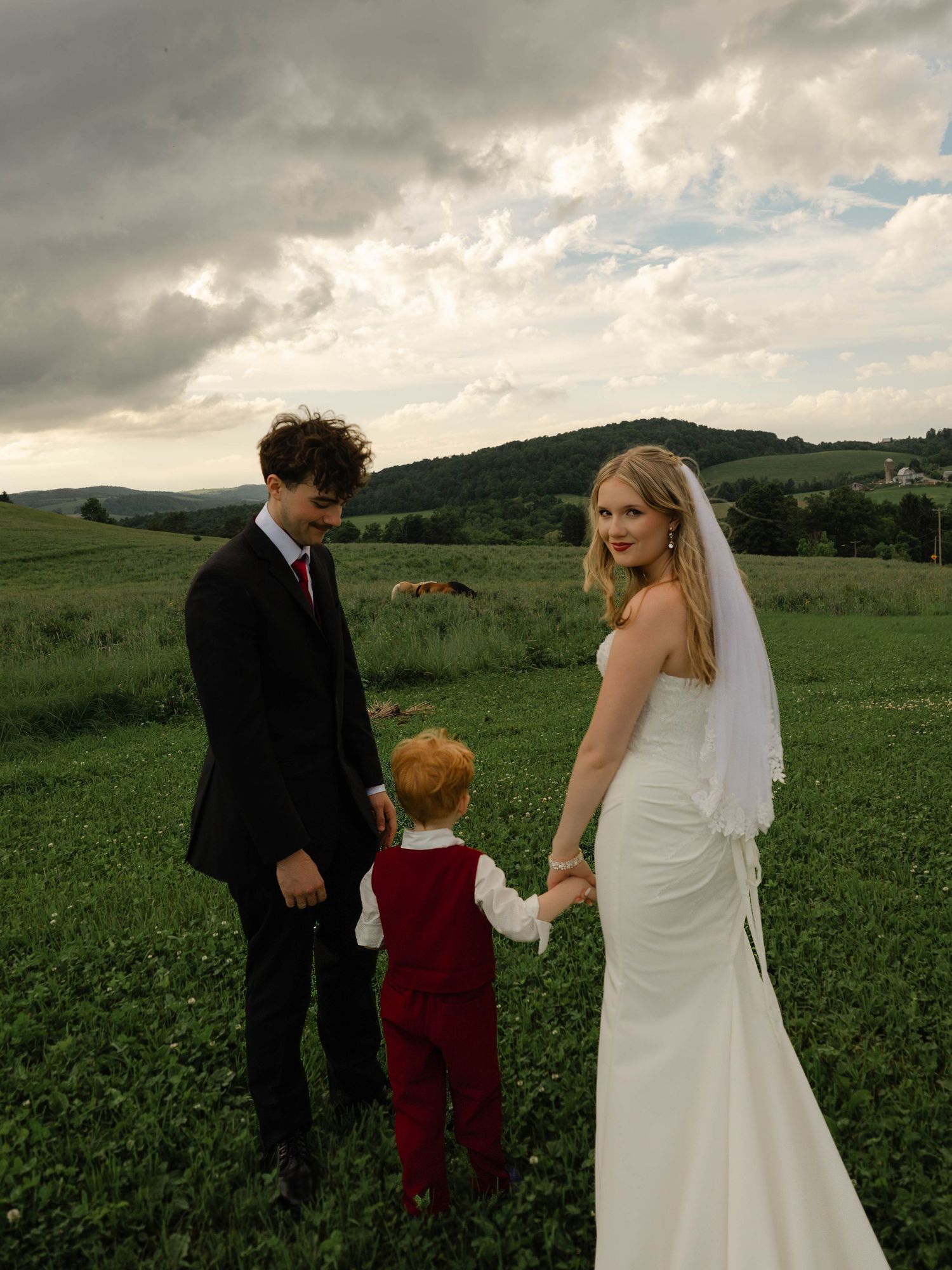 Bride in white gown and groom in black suit hold hands with a young boy in red at an outdoor wedding.
