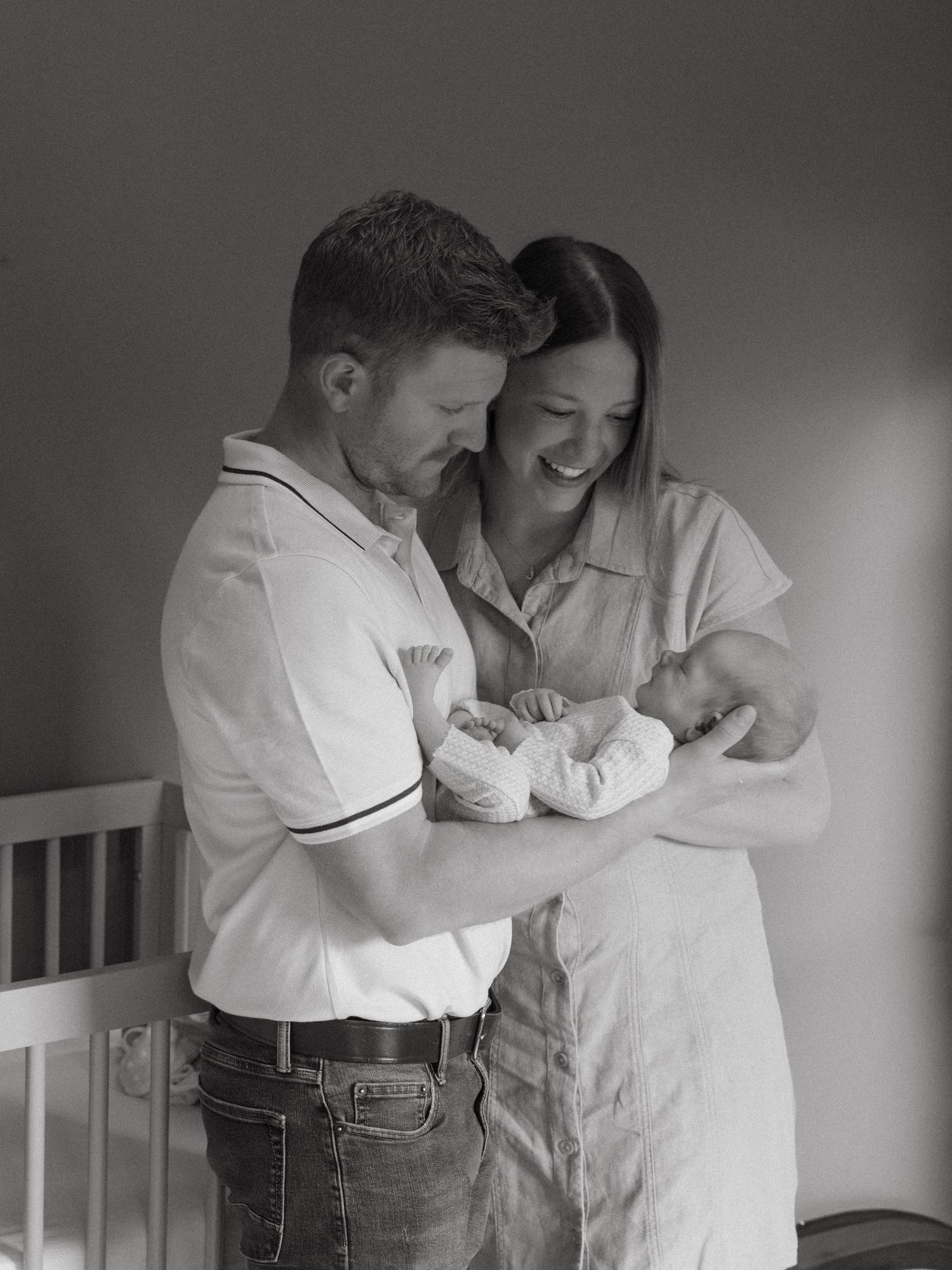 Black and white photo of smiling parents holding their newborn baby together in a nursery room.