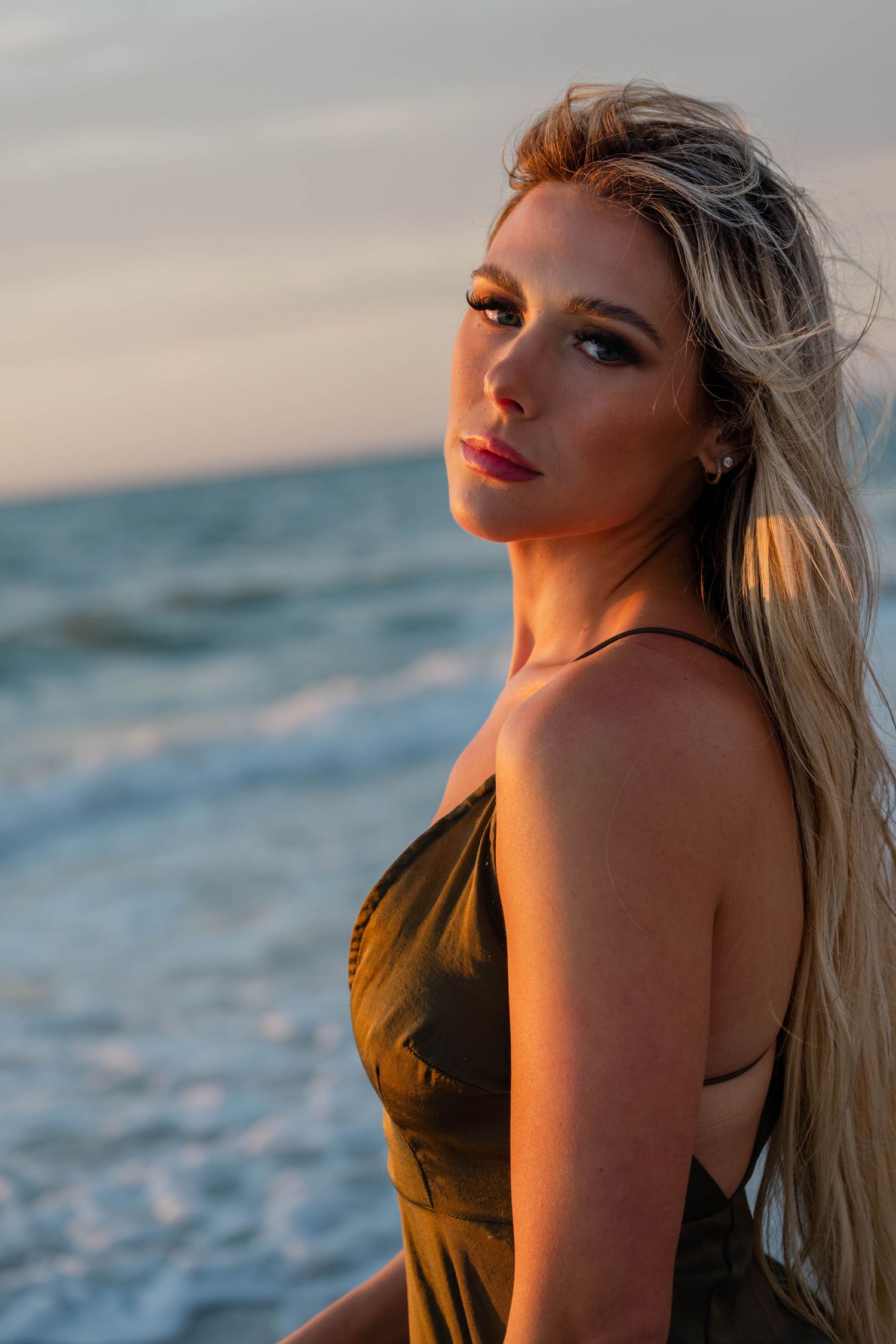 Blonde woman in olive satin dress posing by ocean waves at sunset.