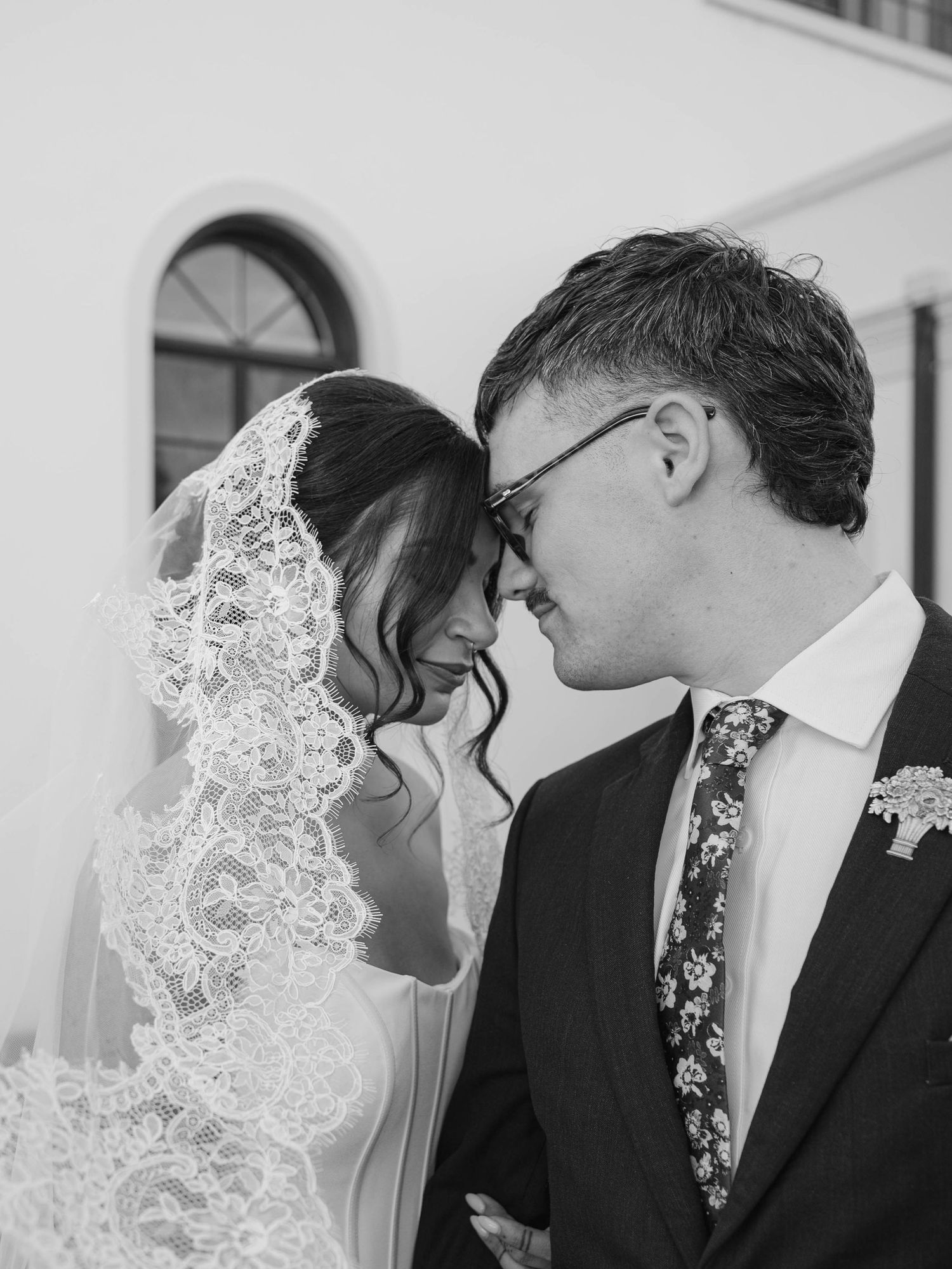 Black and white close-up of bride and groom touching foreheads, bride wearing intricate lace veil.