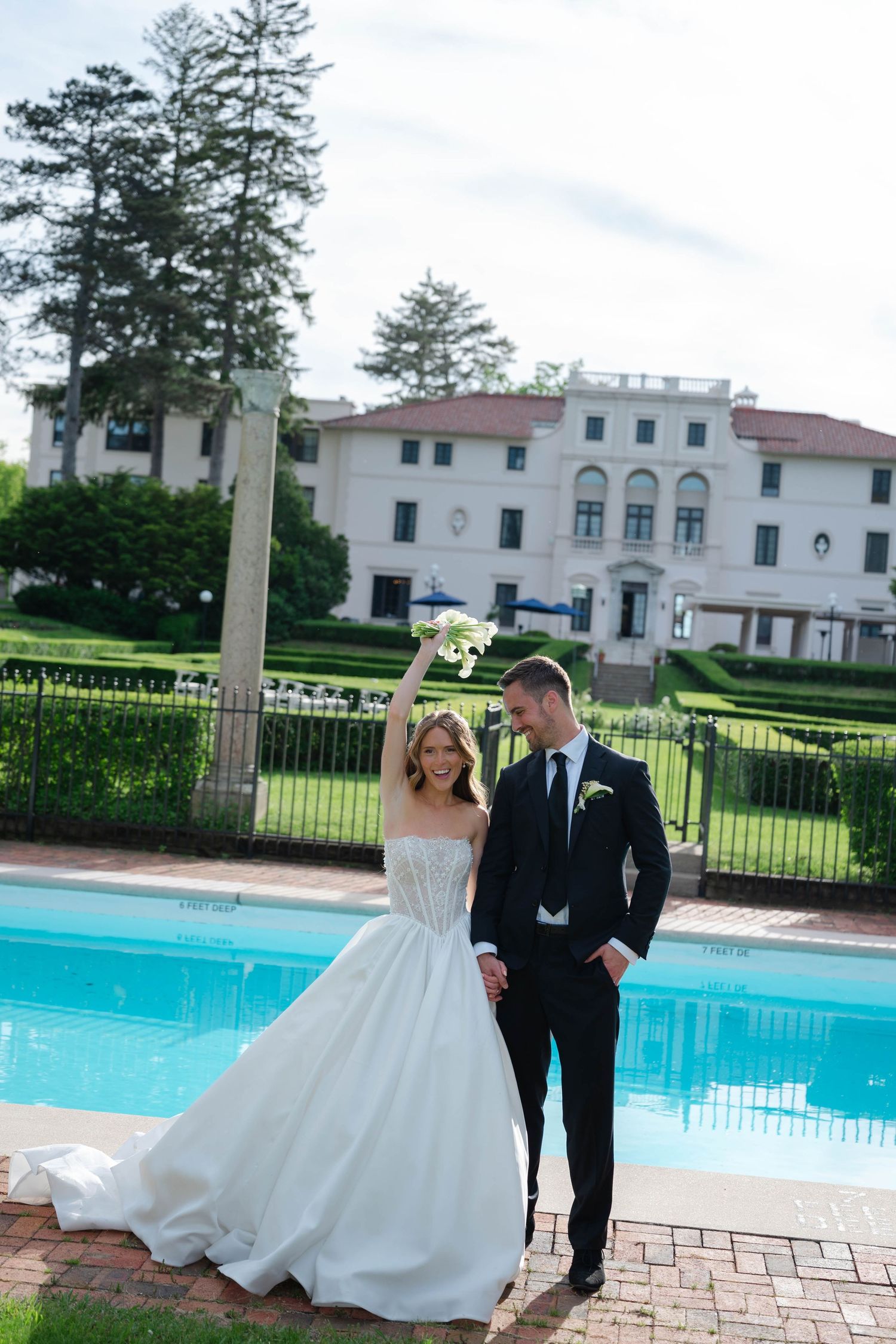 Joyful bride raises bouquet of white calla lilies while groom holds her hand by a pool at a mansion.