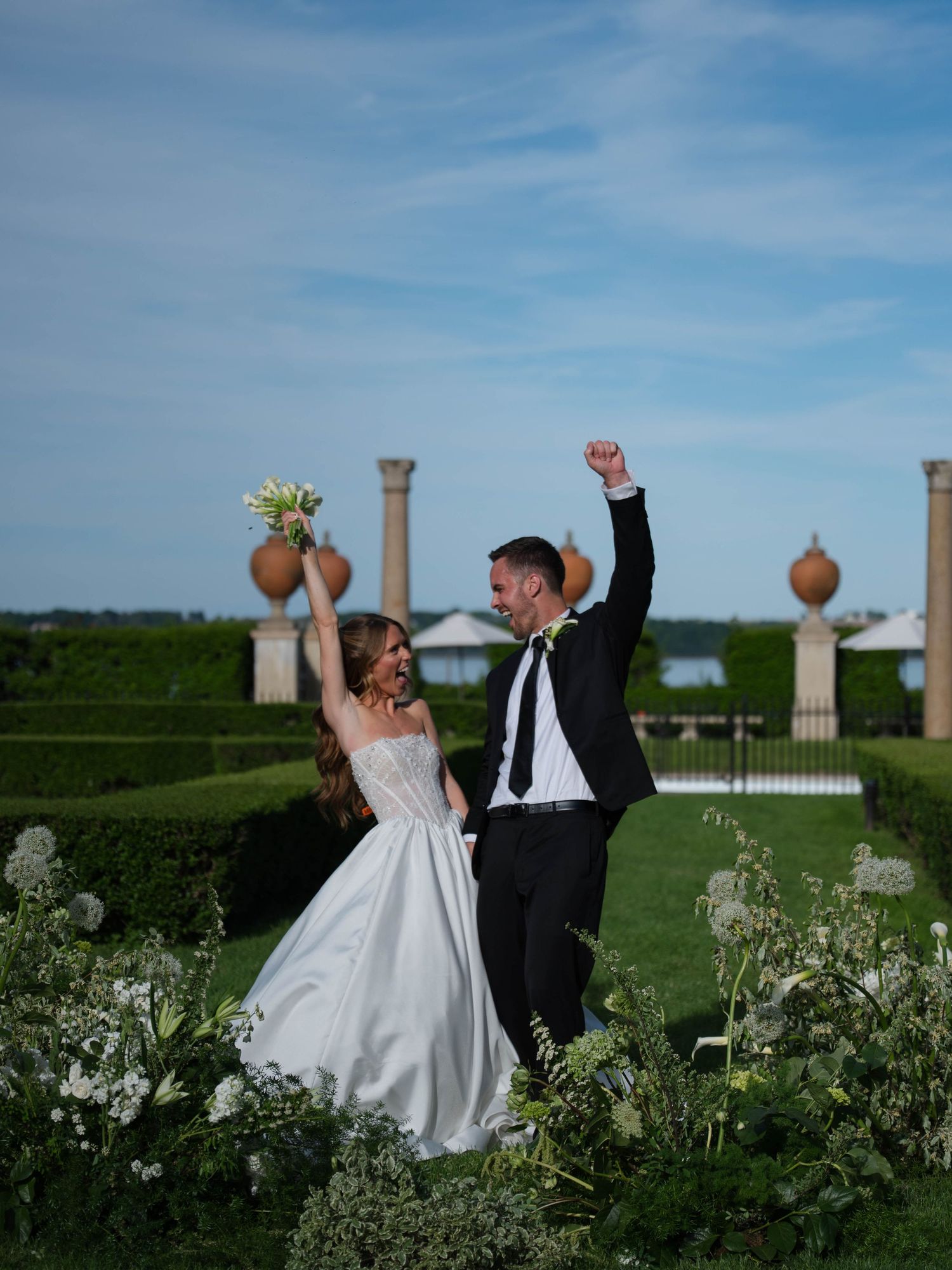 Excited newlyweds cheering with raised arms in lush formal garden with ornamental columns behind them.