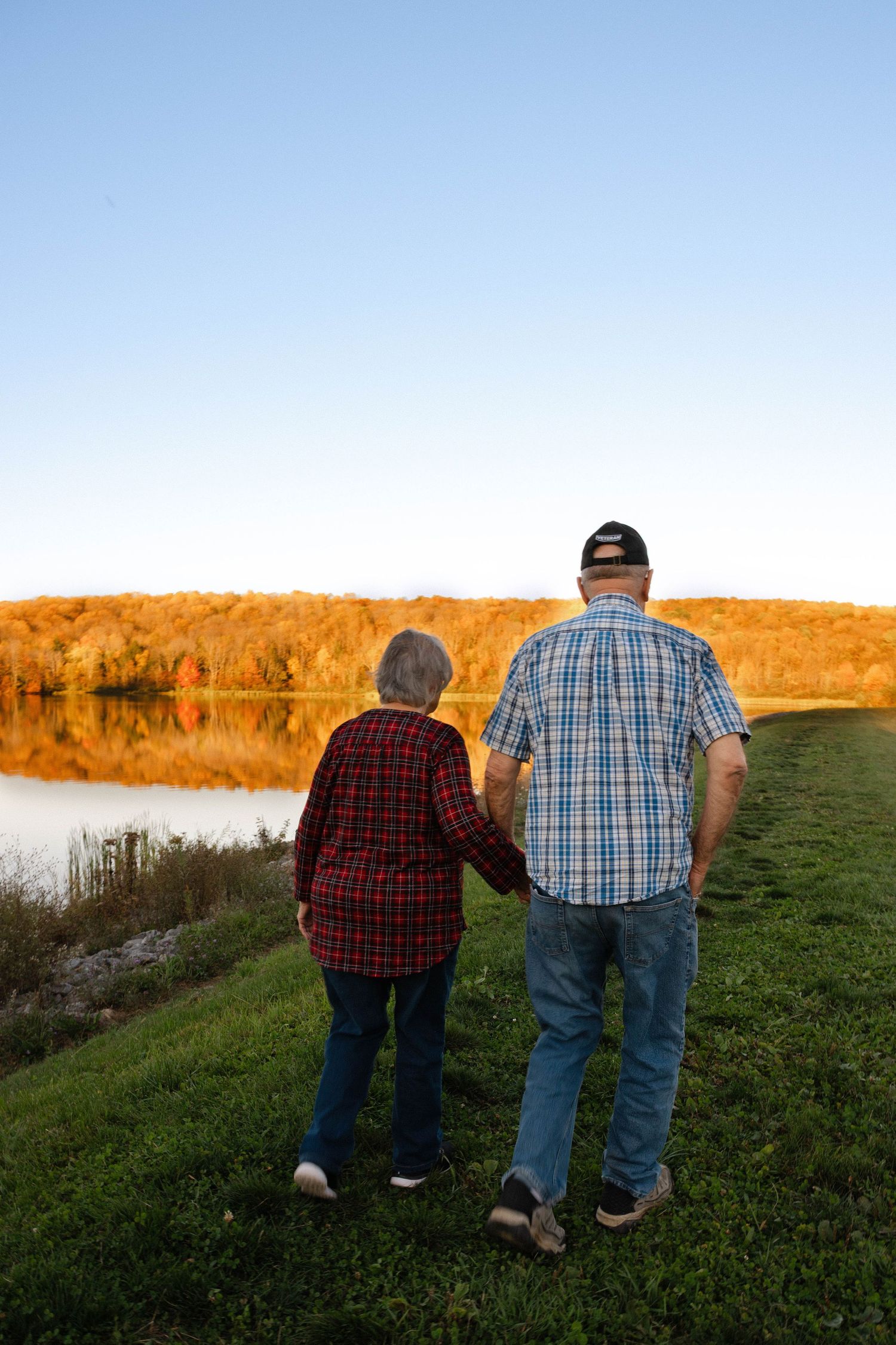 Senior couple holding hands while viewing a calm lake with vibrant autumn foliage reflections.