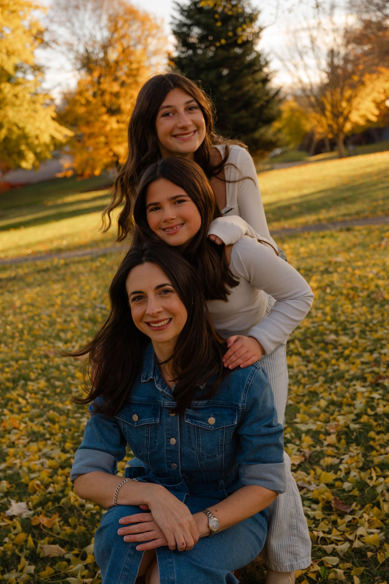Three brunette women pose playfully in a park during autumn, surrounded by golden fallen leaves and colorful trees.
