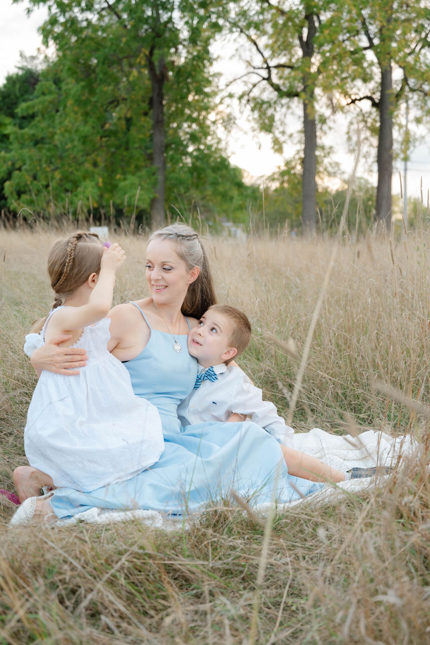 Mother in light blue dress sitting in tall golden grass with two young children during a warm sunset outdoor session.