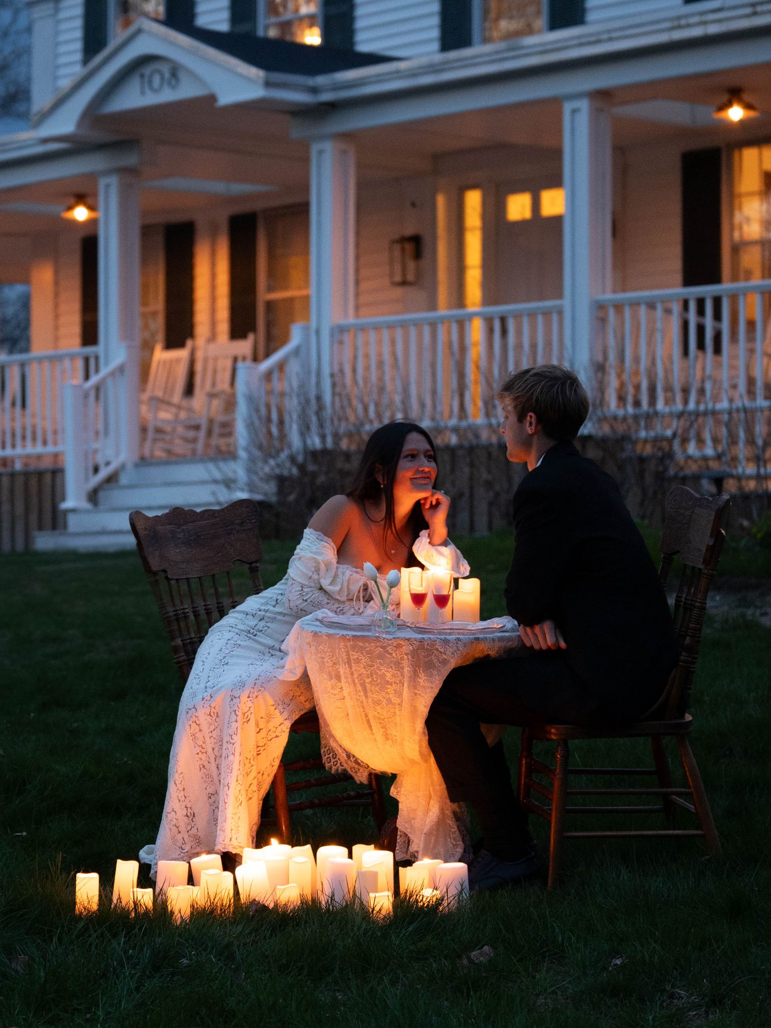 Couple sharing a romantic candlelit dinner outdoors at dusk, surrounded by glowing candles on a lawn.