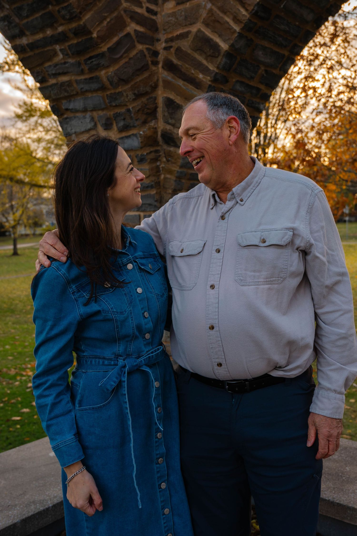 Laughing woman in denim dress and older man share a joyful moment under a stone arch at sunset.