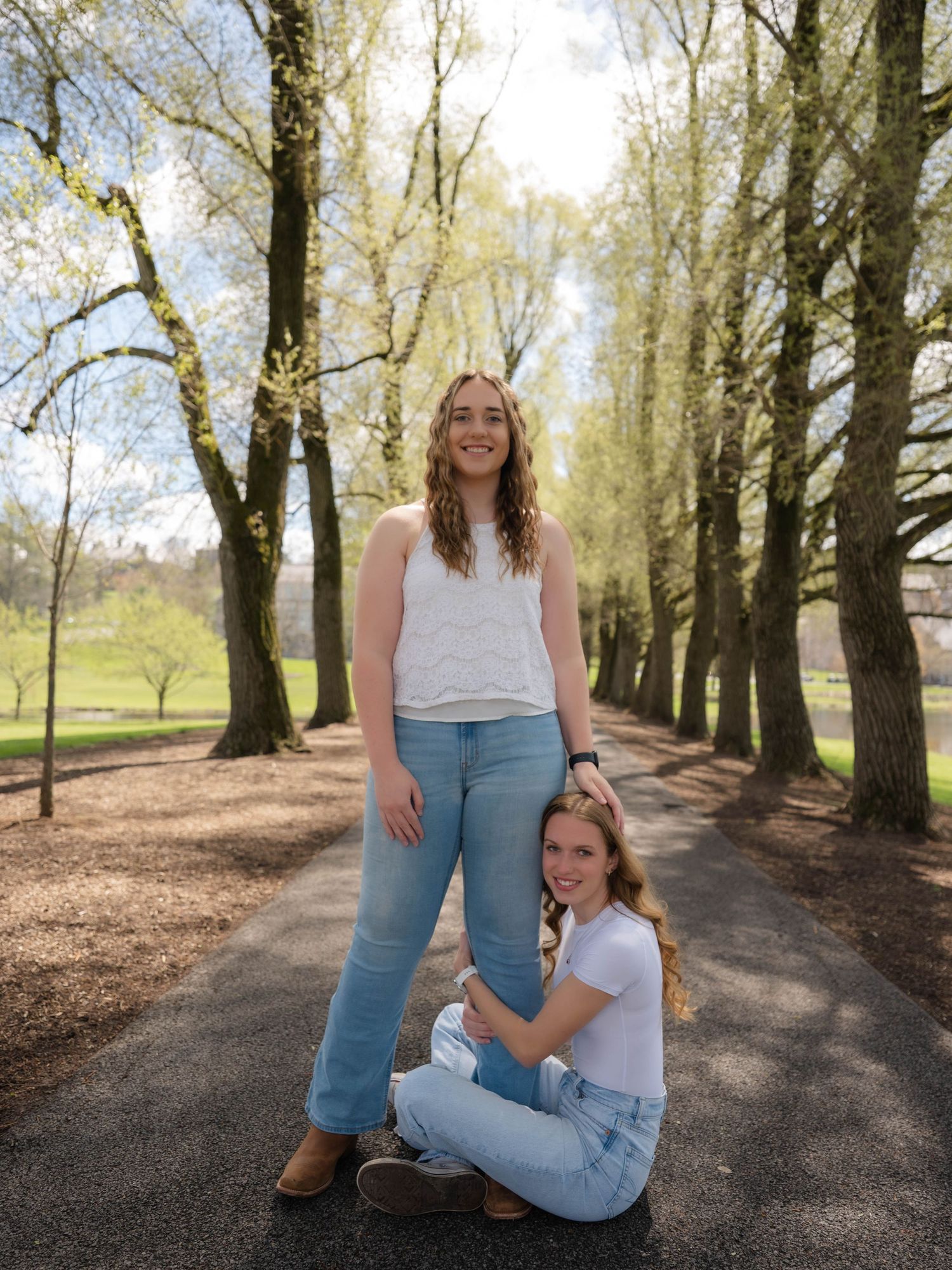 Two young women posing on a tree-lined path in a park during spring, wearing casual jeans outfits.