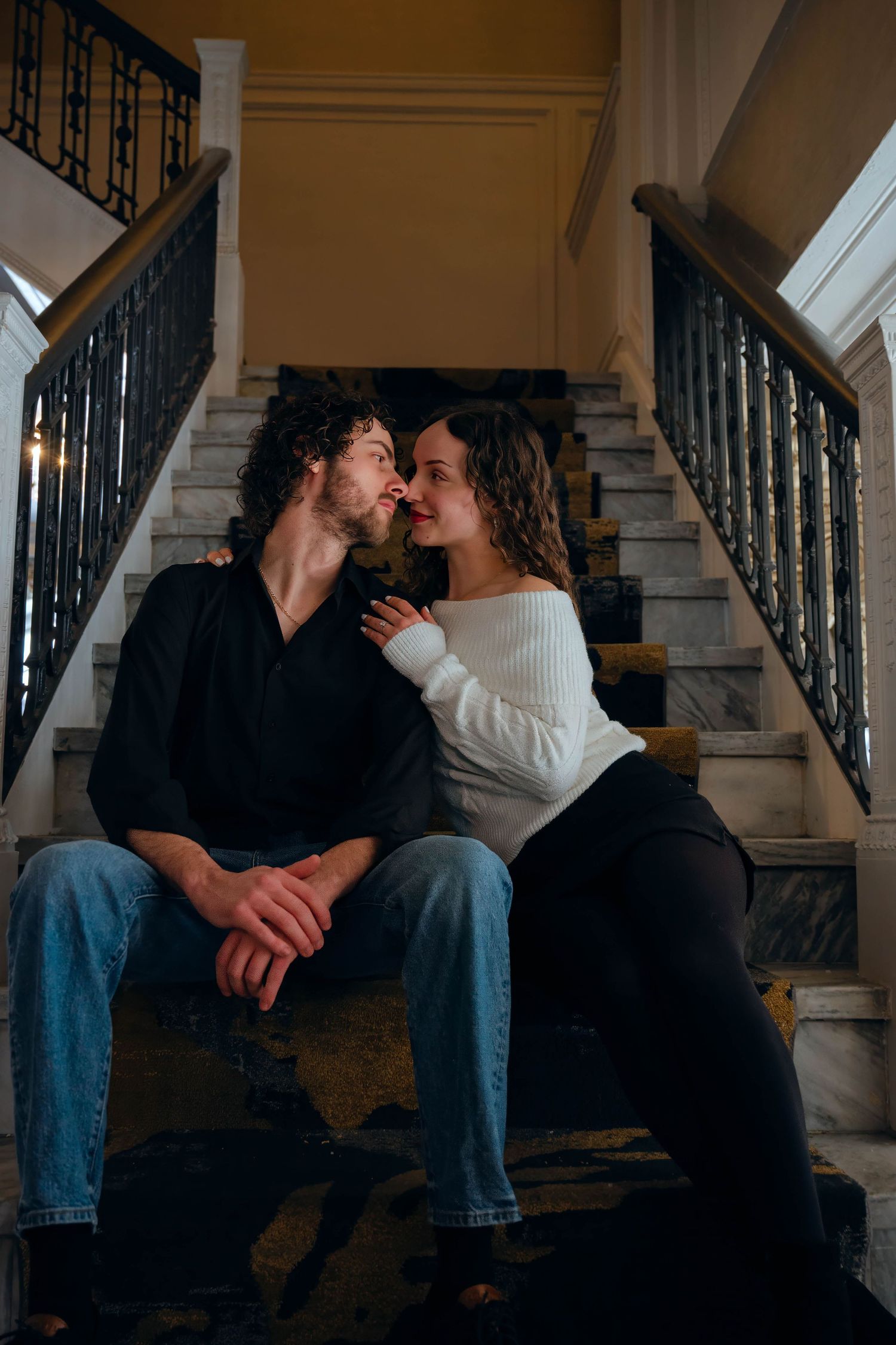 A romantic couple sits close together on elegant marble stairs, sharing an intimate moment in a grand interior.