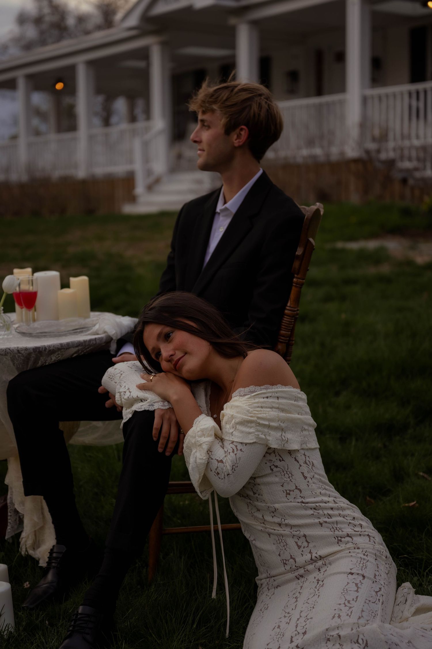 Couple in wedding attire outdoors at dusk, woman in lace dress resting on man's lap near candlelit table. Maine Wedding