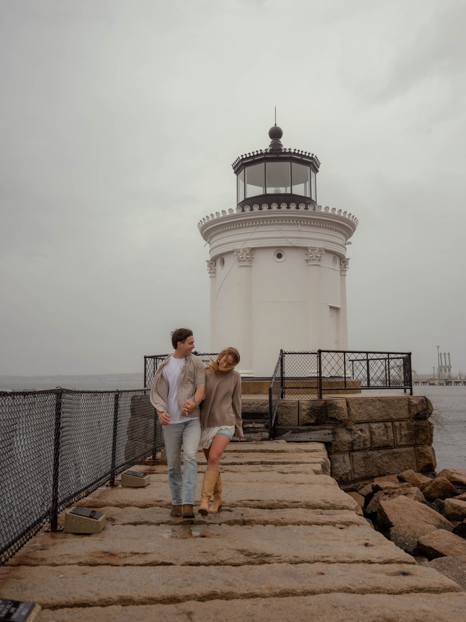 Happy couple walking hand in hand along stone path beside white lighthouse.