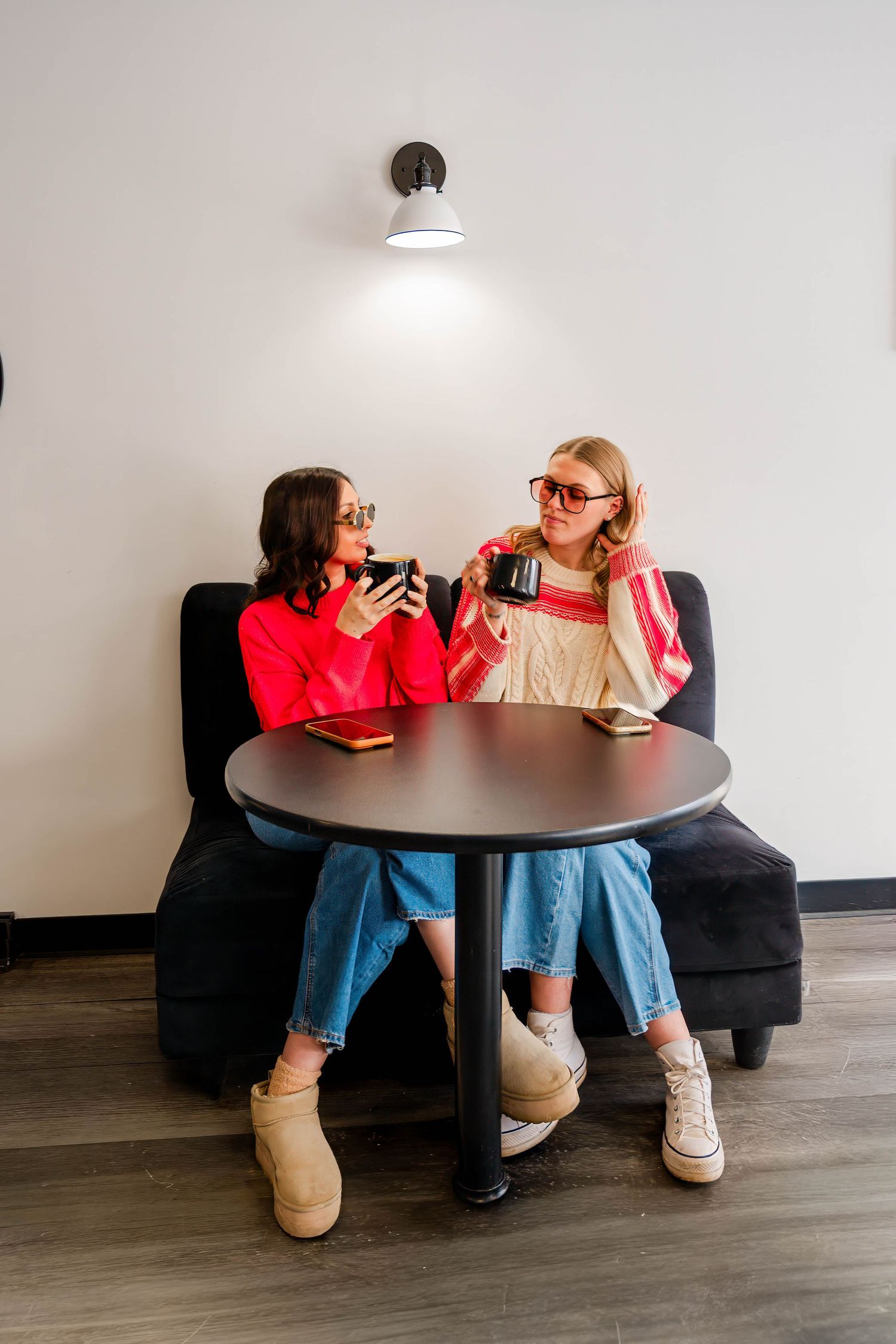 Two women sitting at a round table in a cafe, chatting and holding coffee mugs.