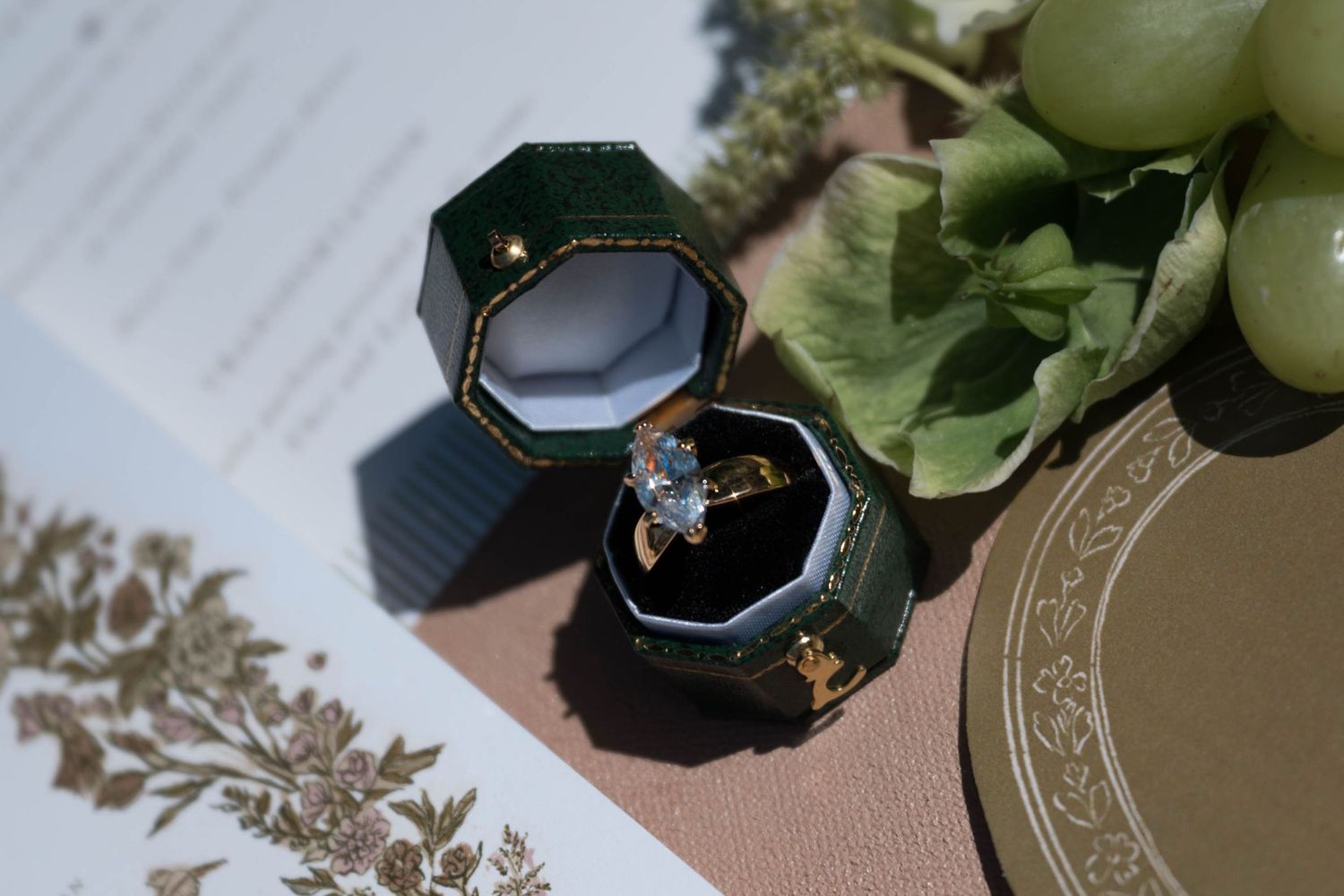Diamond engagement ring displayed in a green velvet hexagonal ring box with floral stationery backdrop.