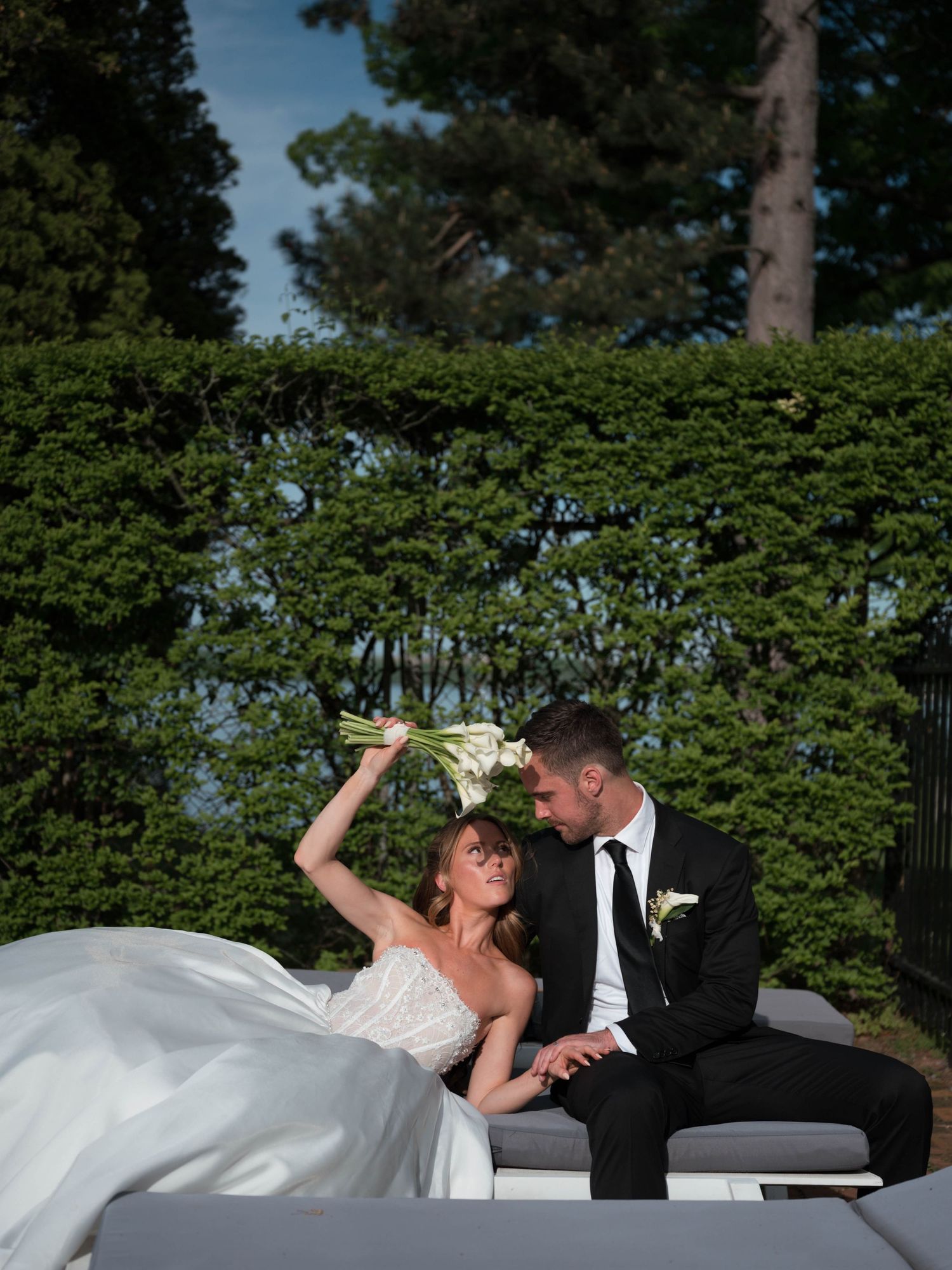 Bride playfully raises white calla lily bouquet while groom gazes at her on outdoor lounge.