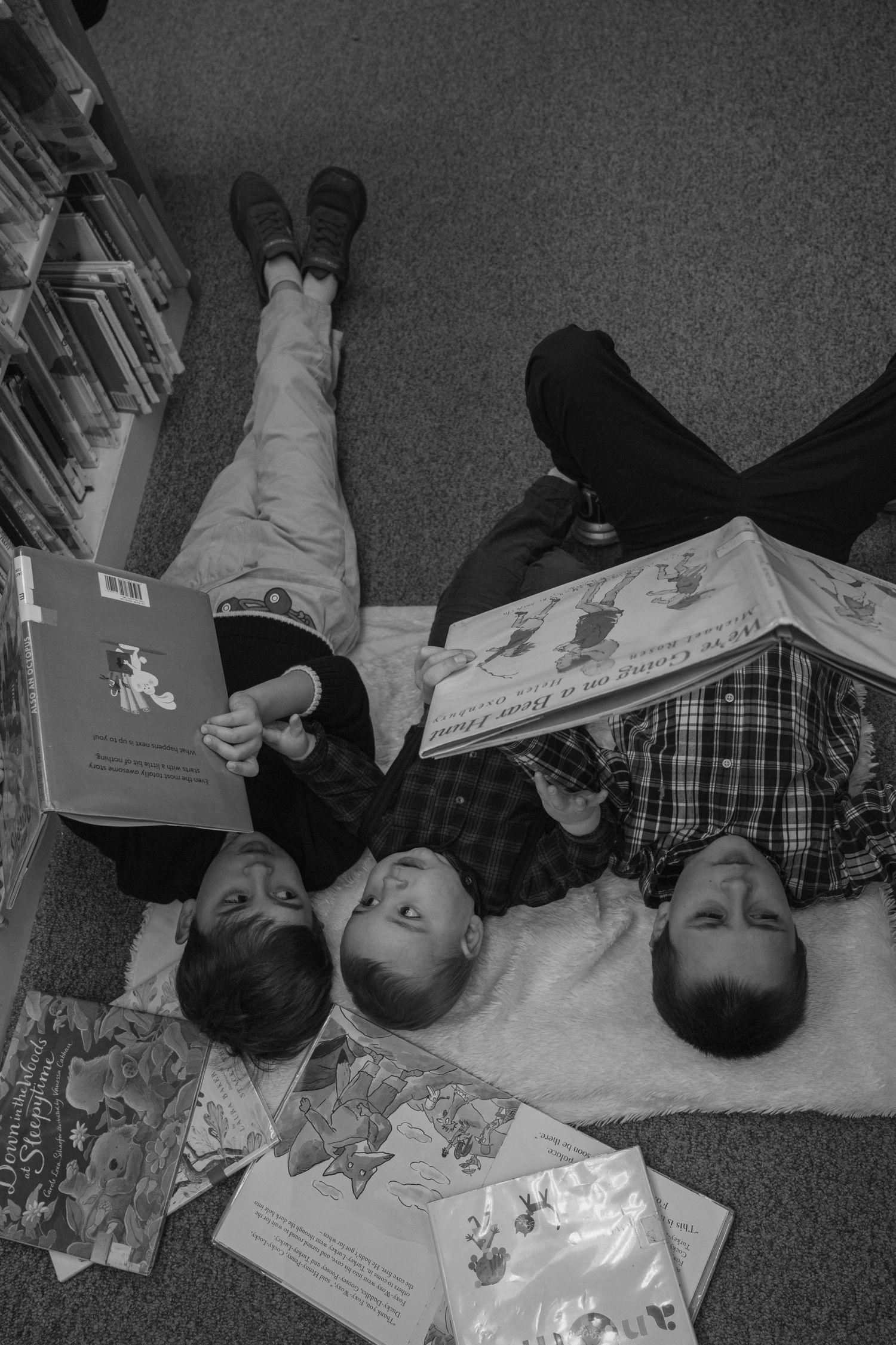 Overhead black and white photo of three siblings lying on floor reading books in a library.